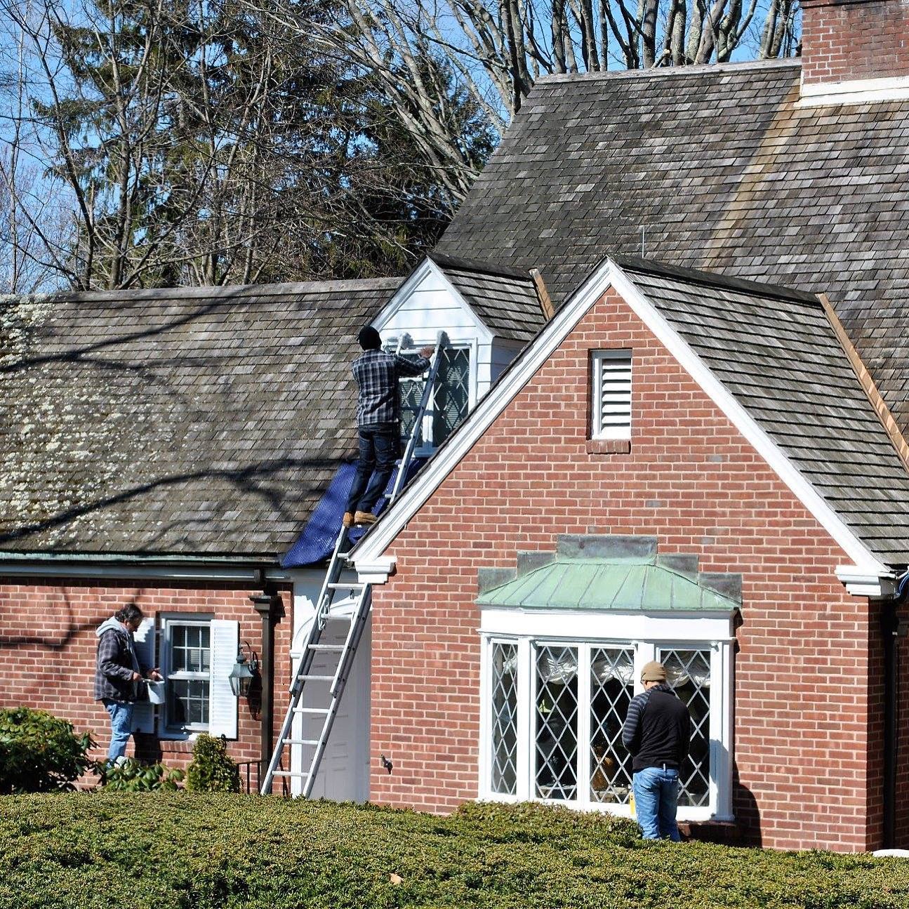 Workers on a brick house with a weathered roof. One on a ladder near a small window, others near windows on the ground level.