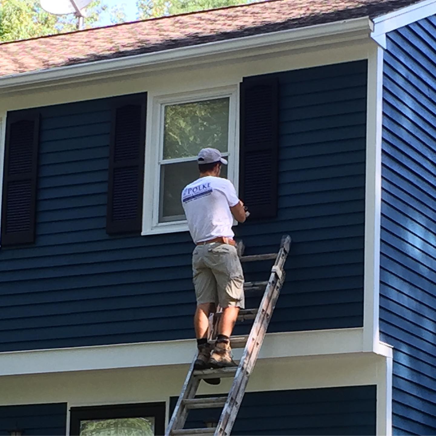 Person on a ladder painting a blue house with black shutters. He is wearing a white shirt and khaki shorts.