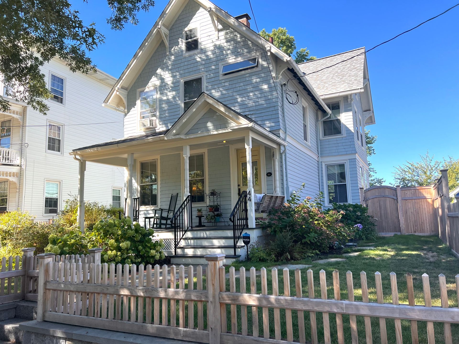 Light blue house with a porch and picket fence on a sunny day.  Black chairs on the porch.