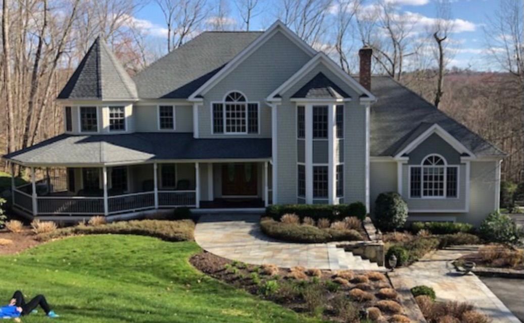 Two-story light green house with a wraparound porch and manicured landscaping. A person is sitting on the front lawn.