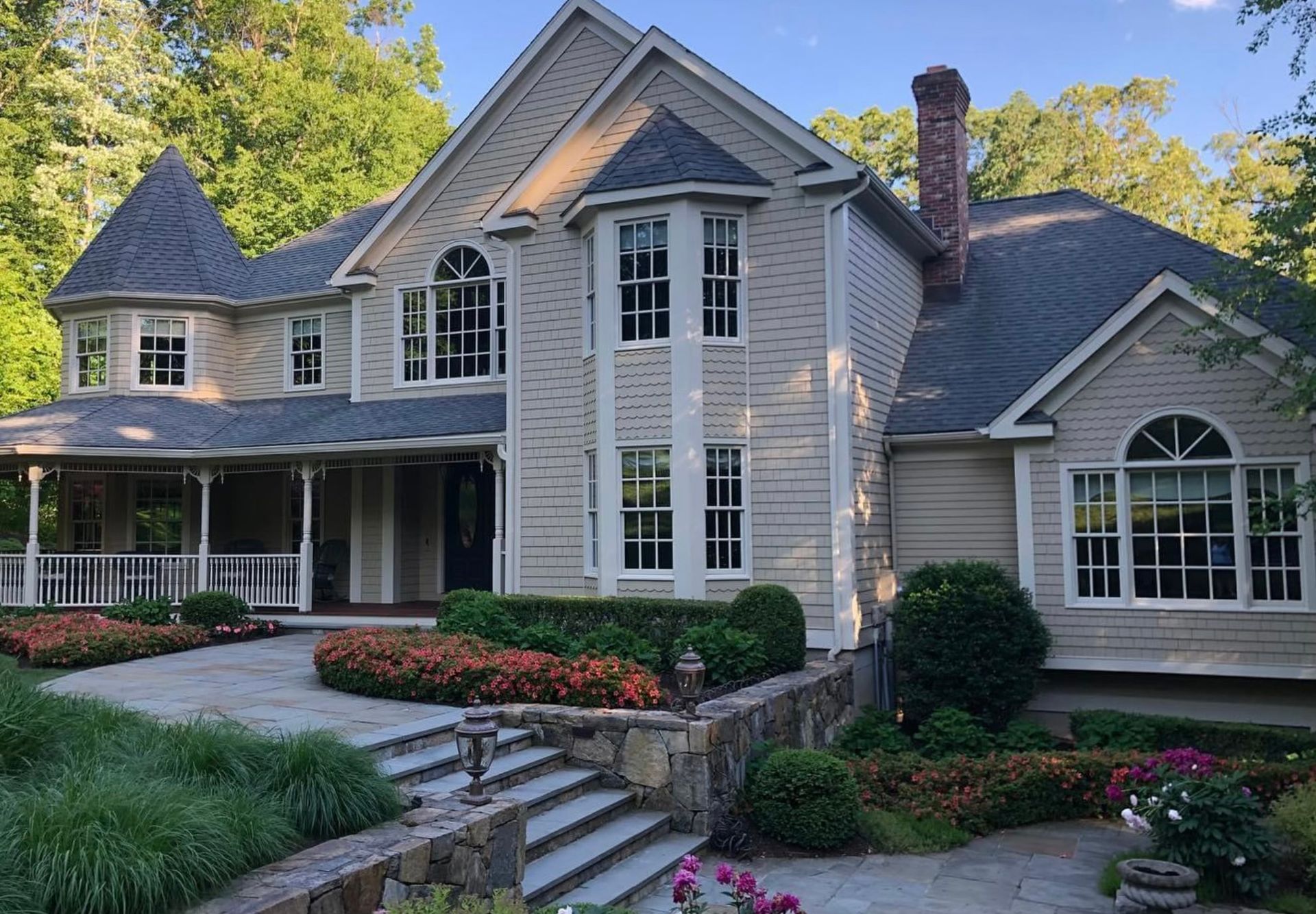 Beige two-story house with multiple windows, a turret, and a porch, set in a yard with stone steps and flower beds.
