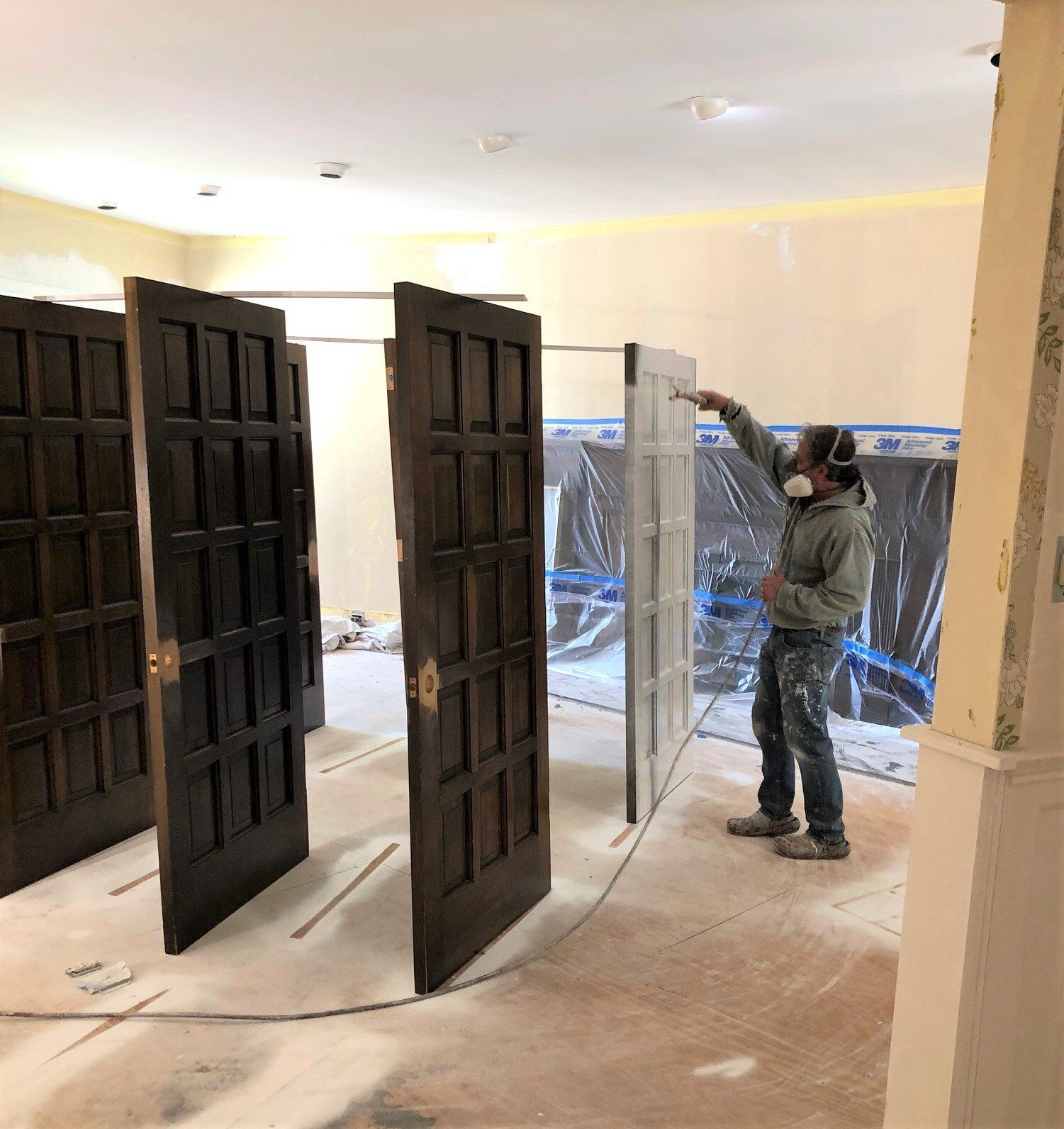 A person spray painting a door white, with other dark doors nearby. They're in a room with protective coverings and wearing a respirator.