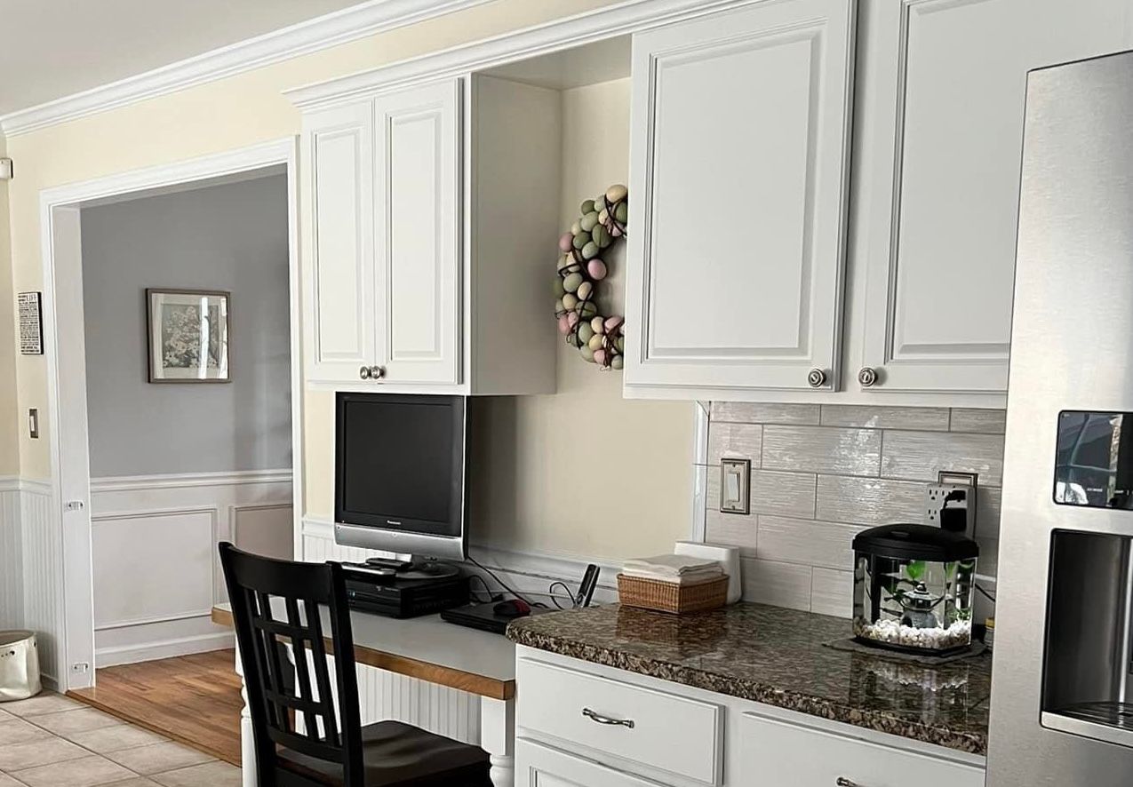 A kitchen with white cabinets, granite countertop, and a built-in desk area. A black chair sits at the desk with a computer.