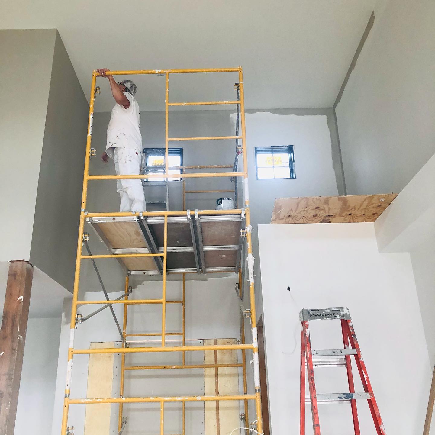 A person paints a high ceiling from a yellow scaffold. They are in a mostly white room with a gray wall. A red ladder is visible.