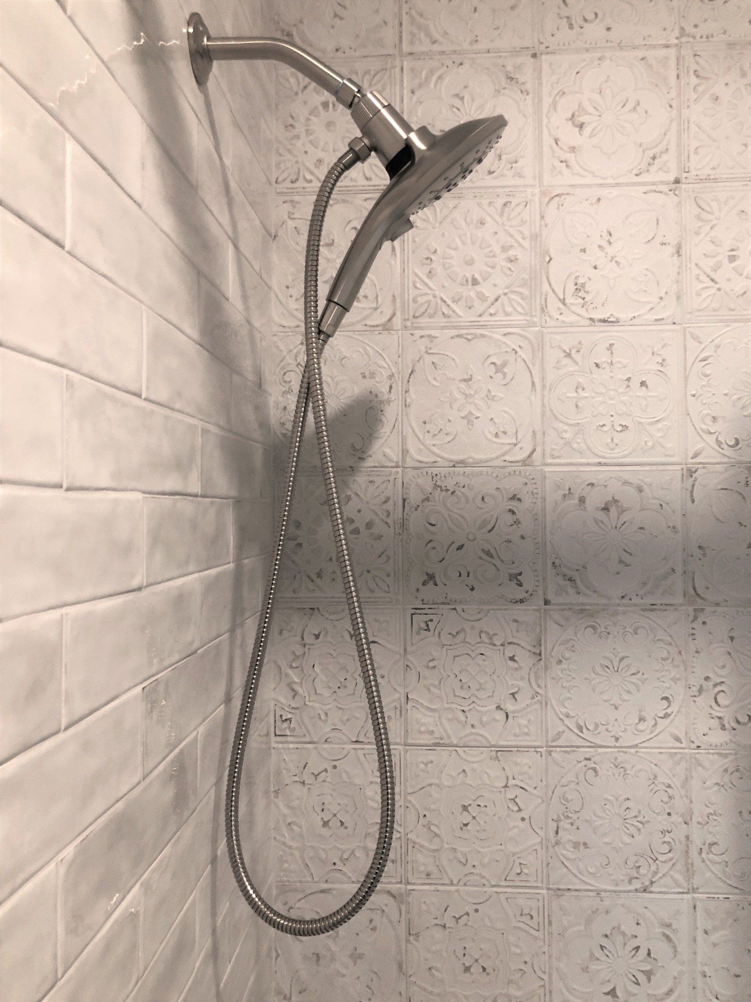 Shower with a handheld showerhead and a silver hose against a white tiled wall.