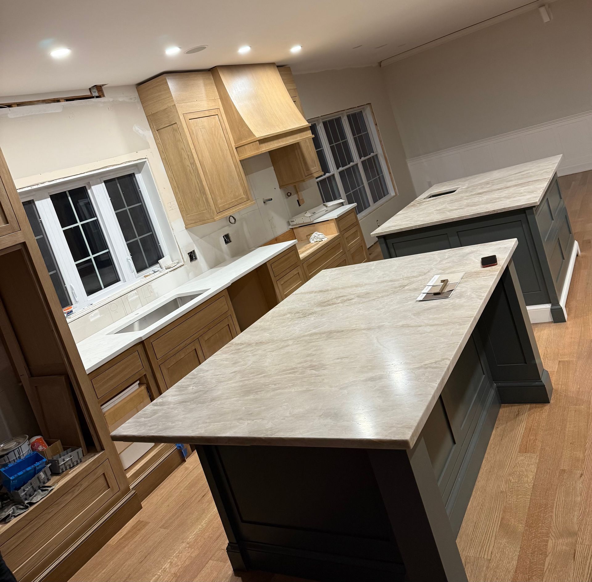 Bright kitchen with two large island tables, light wood cabinets, white counters, and windows at the back.
