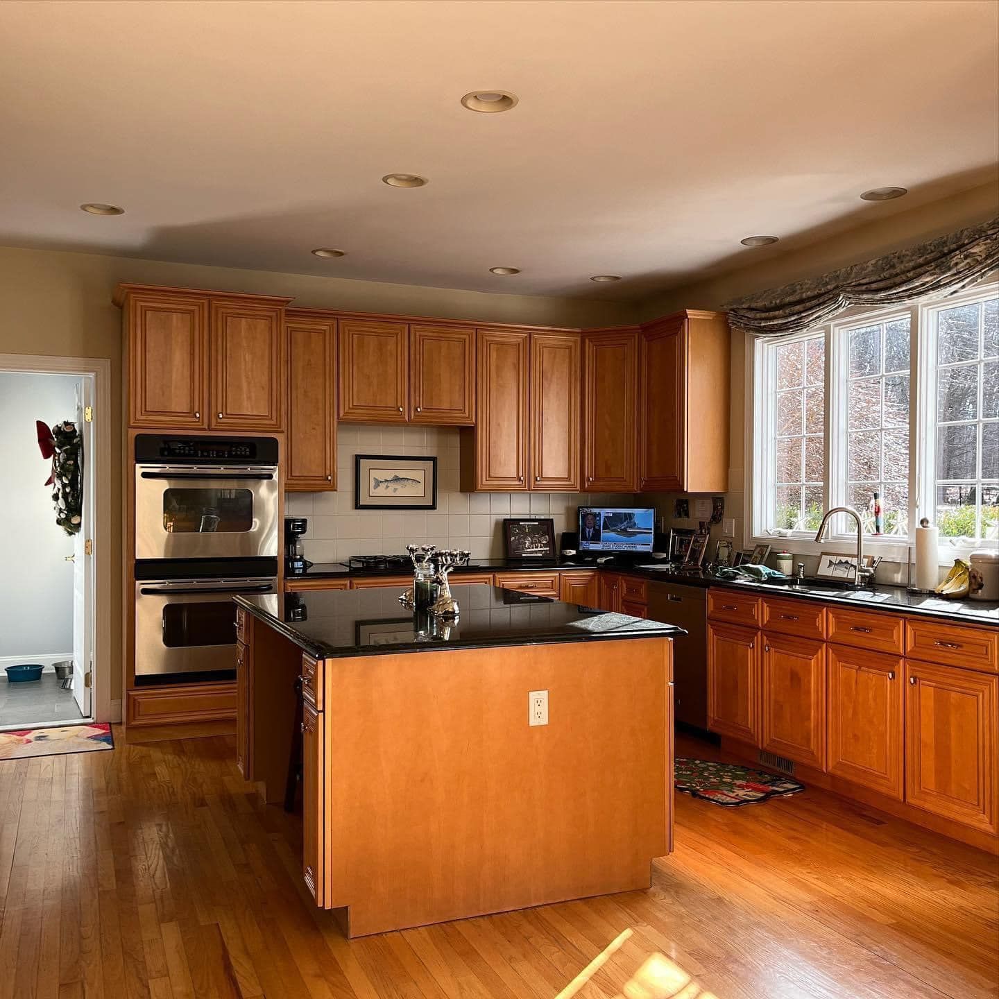 A kitchen with wood cabinets, a black island countertop, stainless steel double ovens, and large windows with a valance.