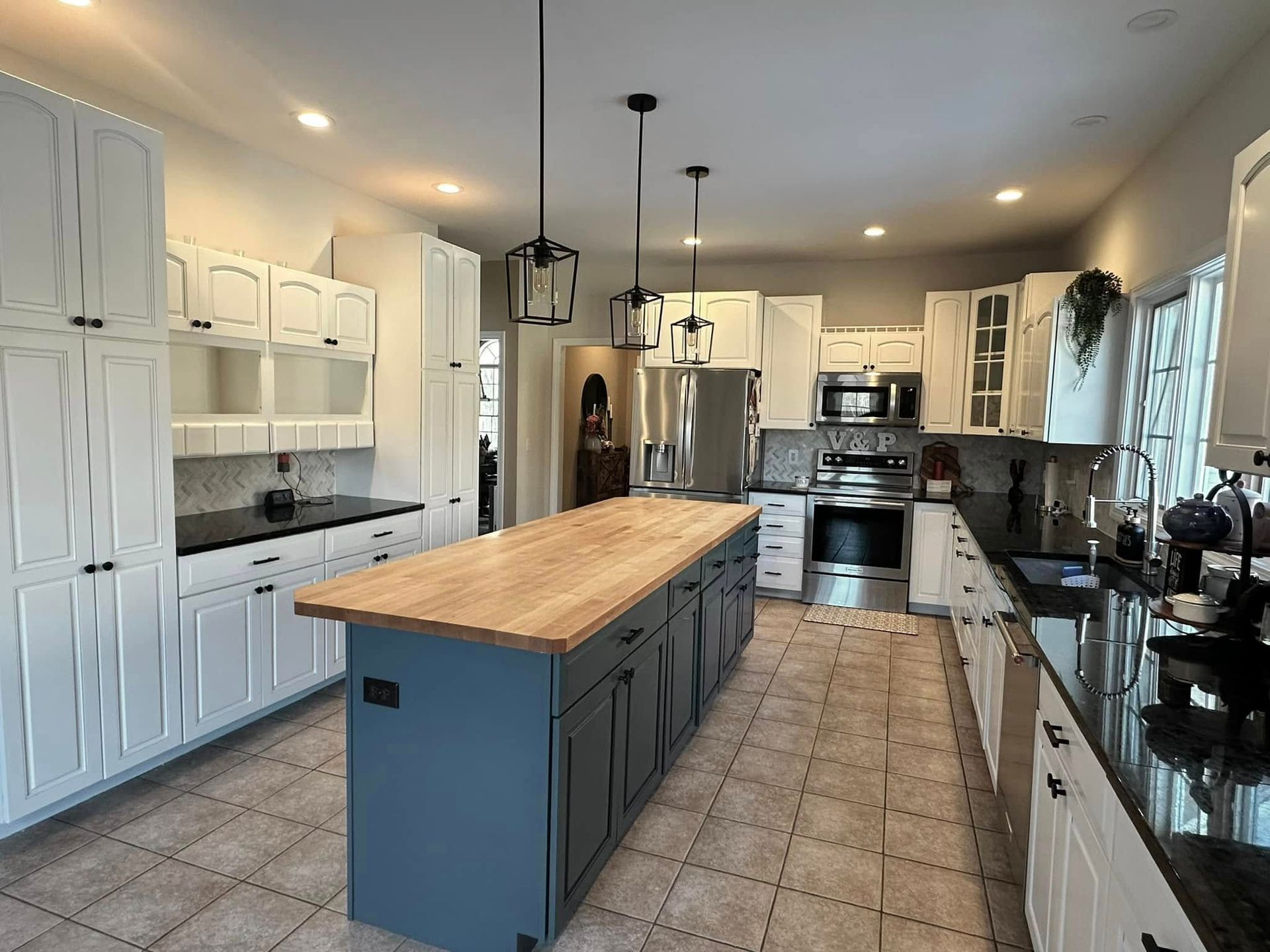 A bright kitchen featuring white cabinets, dark countertops, a large blue island with a wood top, and three pendant lights.