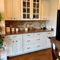 A kitchen buffet with white cabinets, a dark wood countertop, and a brick-patterned backsplash, next to a wood chair.