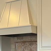 A cream-colored, custom-built kitchen vent hood positioned above a tiled backsplash with a decorative pattern.