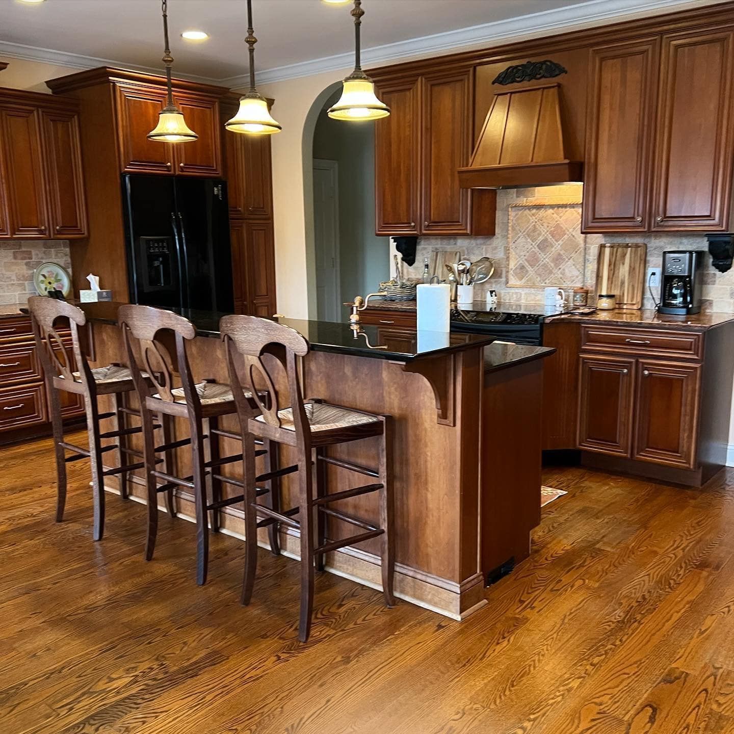 A kitchen with rich wood cabinets, a black refrigerator, a granite-topped island, three wooden bar stools, and wood floors.