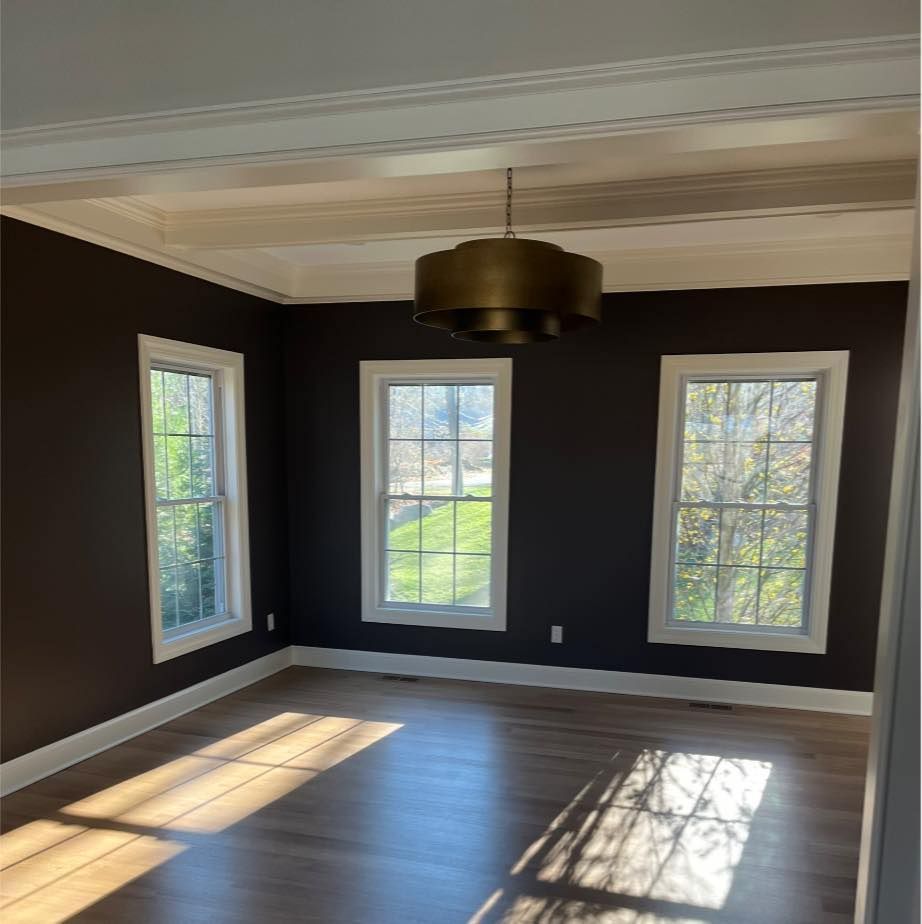 Empty room with dark brown walls, light hardwood floors, three white-trimmed windows, and a large tiered brass chandelier.