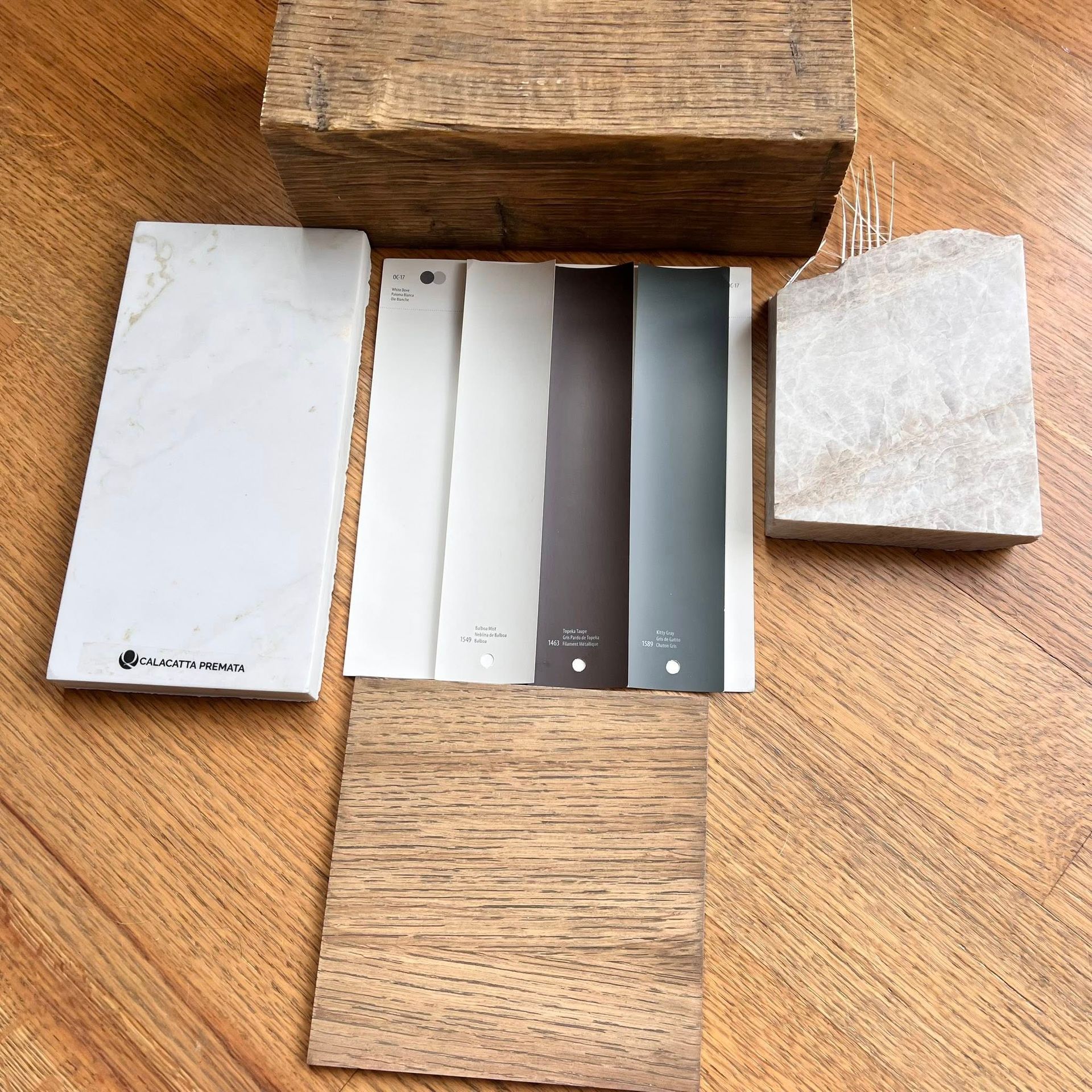 A design materials flat lay featuring wood samples, marble and stone swatches, and a set of grey and brown paint strips.