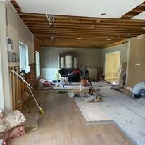 An interior view of a room under renovation with unfinished walls, exposed wooden ceiling beams, and new wood flooring.