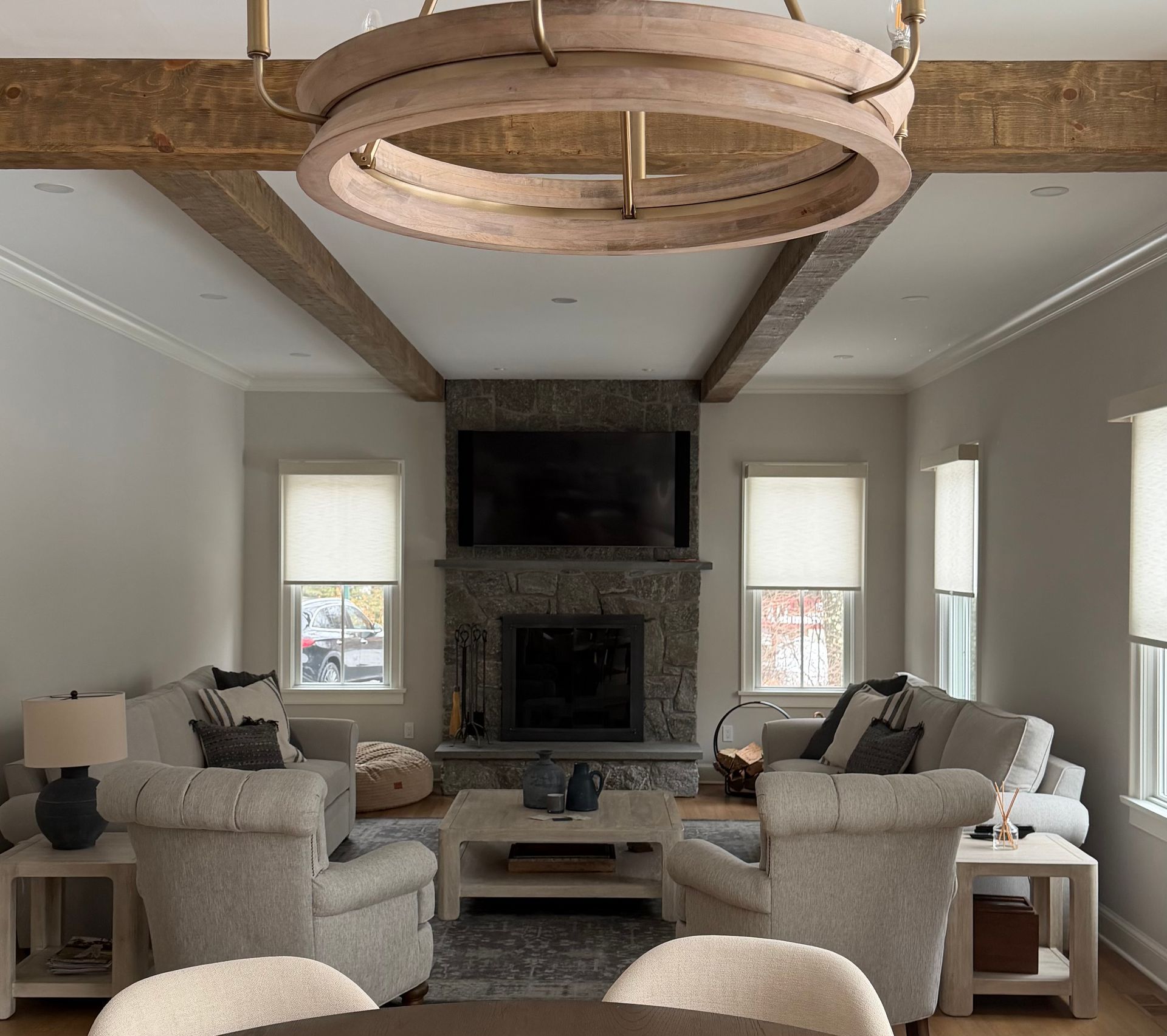 A neutral-toned living room featuring a stone fireplace, exposed wooden ceiling beams, a round chandelier, and plush sofas.