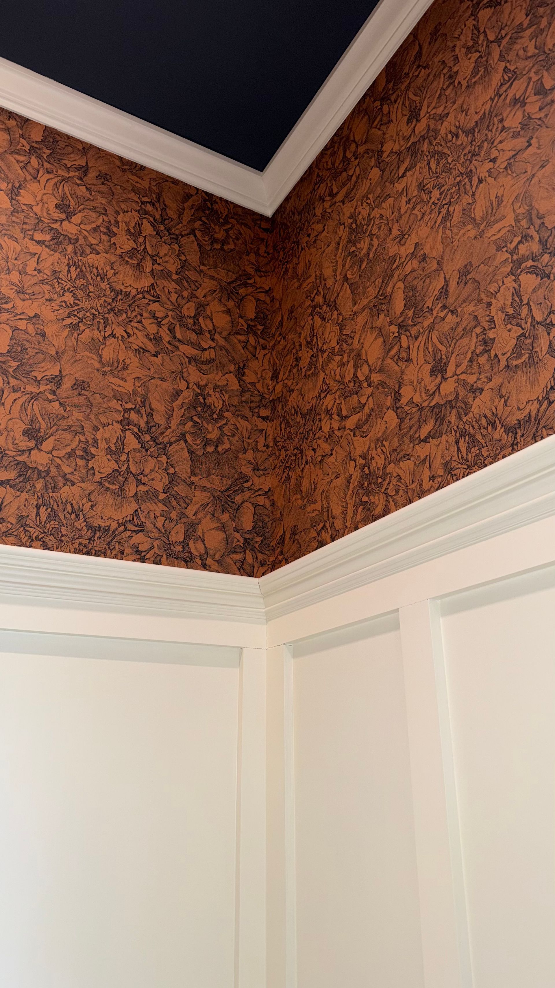 A corner of a room featuring terracotta-colored wallpaper with a dark floral pattern above white wainscoting and a ceiling.