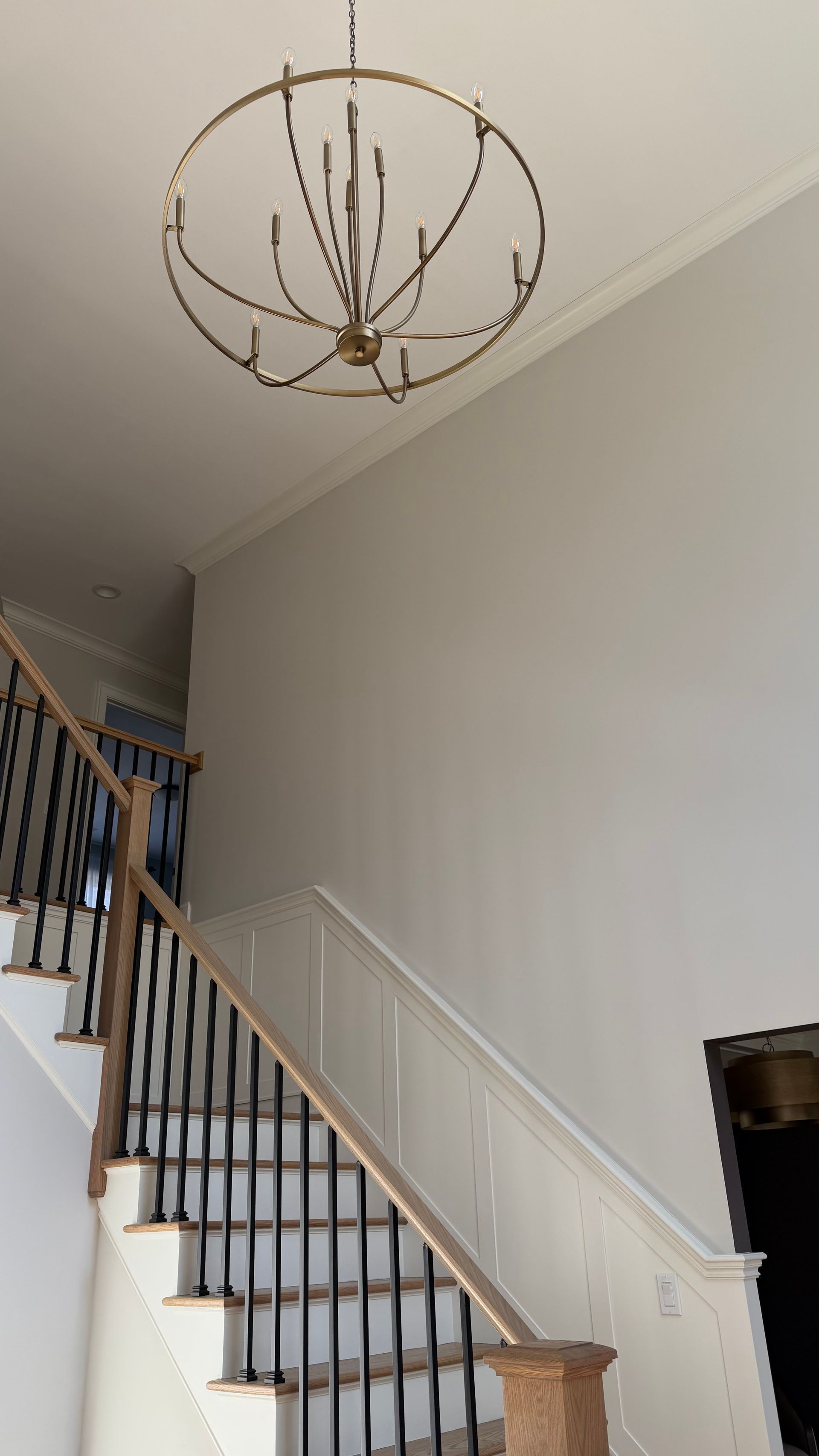 A staircase with black balusters and a wood handrail leads up a tall, light-gray wall under a gold orb-style chandelier.