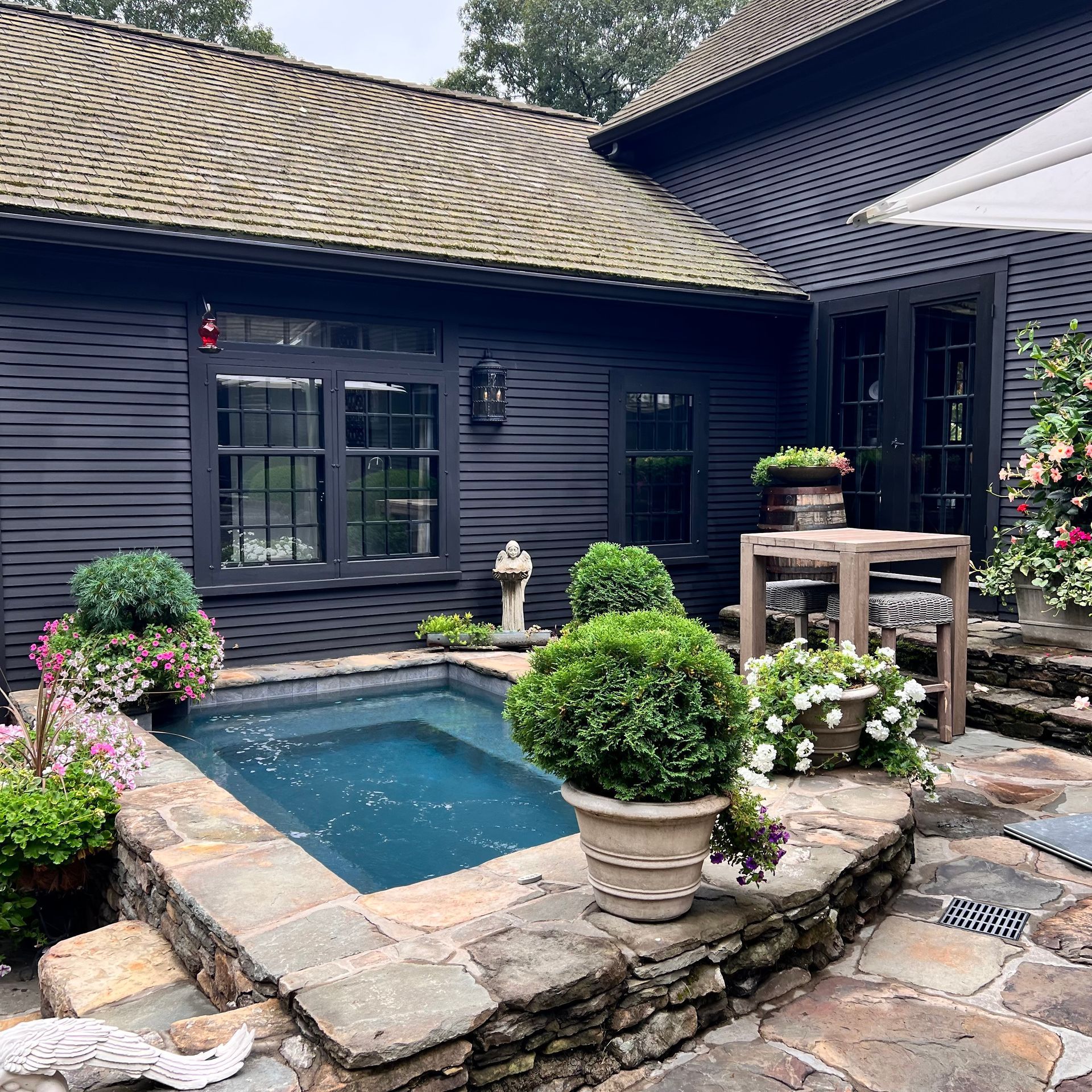 A rectangular stone-lined plunge pool nestled against a dark, wooden-sided house with potted plants and a stone patio.