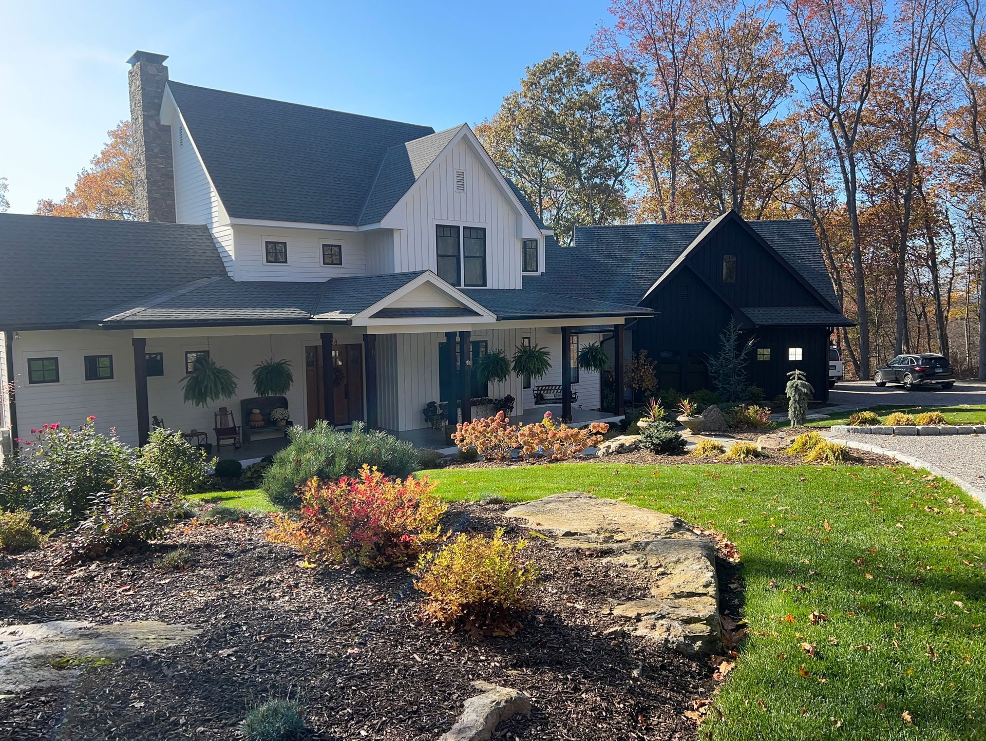 A white, multi-level farmhouse with a black roof and attached dark-colored garage, surrounded by landscaping and trees.