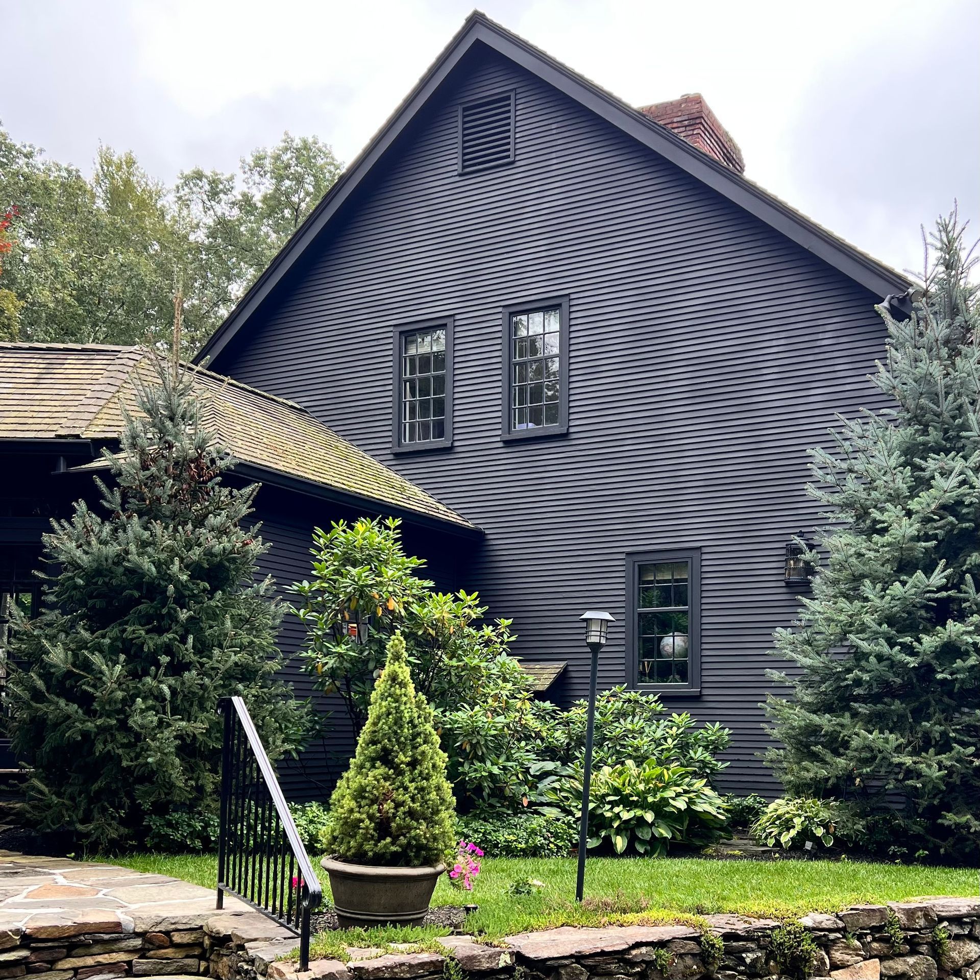 A dark, shingled, two-story house with a gabled roof, nestled among pine trees, shrubs, and a stone retaining wall.
