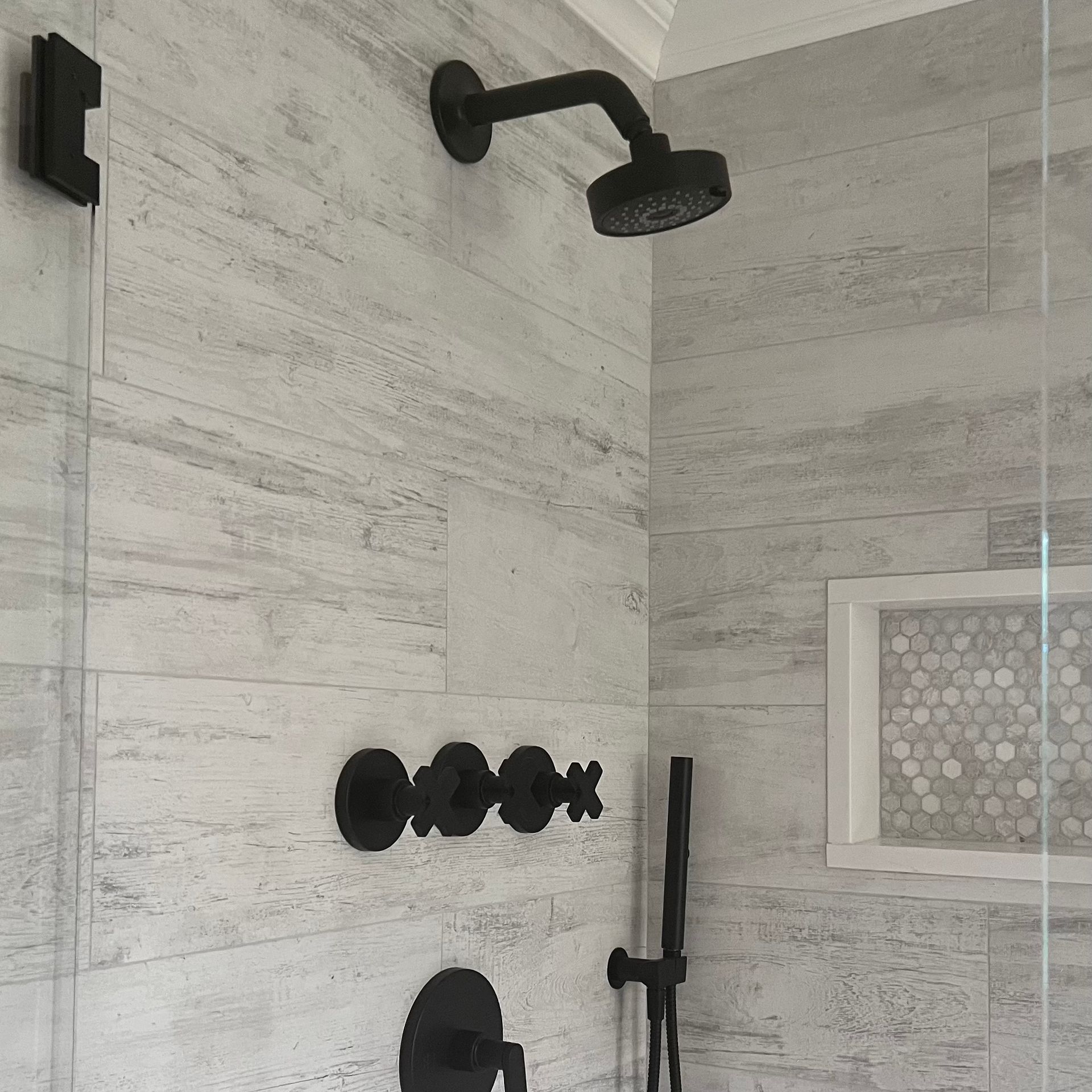 Black shower fixtures against light gray tiled wall. Includes showerhead, faucet controls, handheld shower, and accent mosaic.