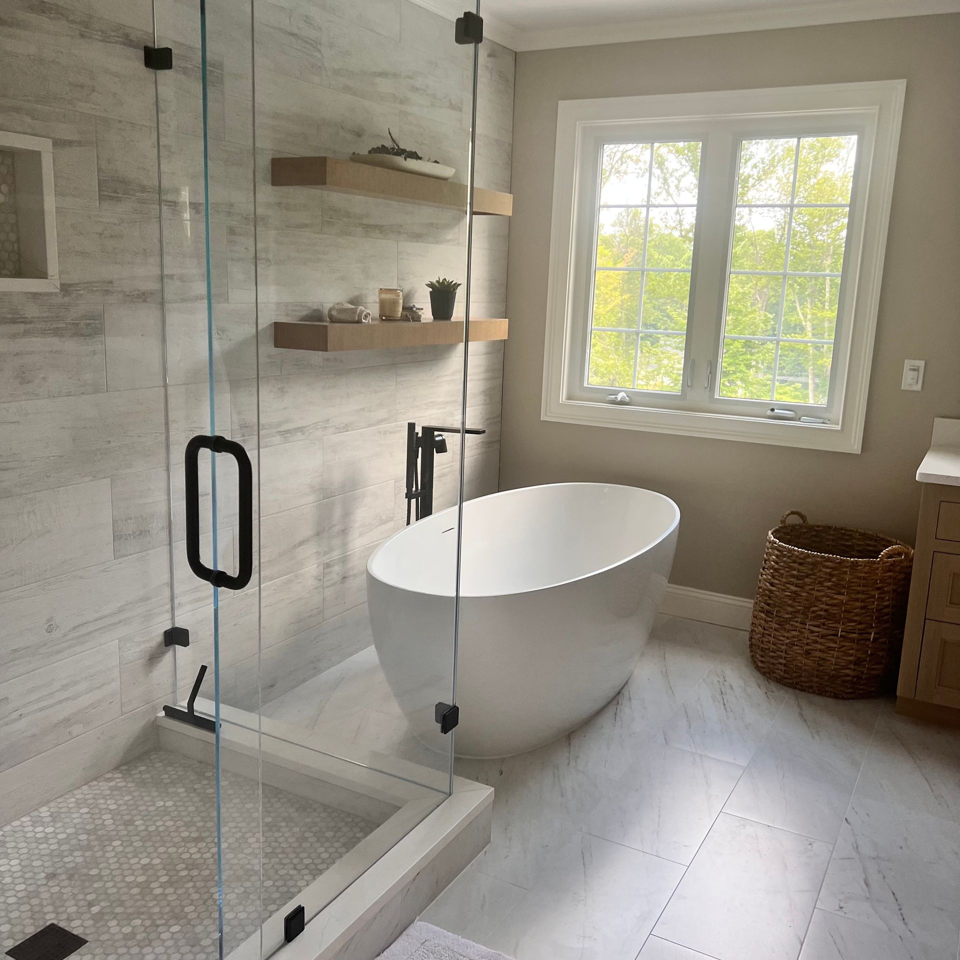 Modern bathroom with a glass-enclosed shower, freestanding tub, and a window overlooking trees.
