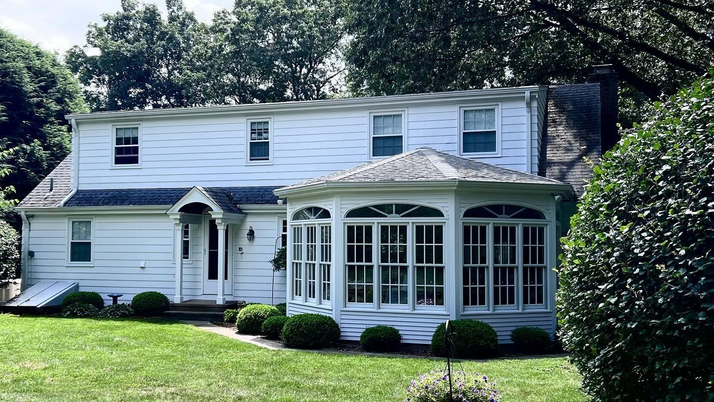 White two-story house with an attached sunroom and green lawn. Overcast day.