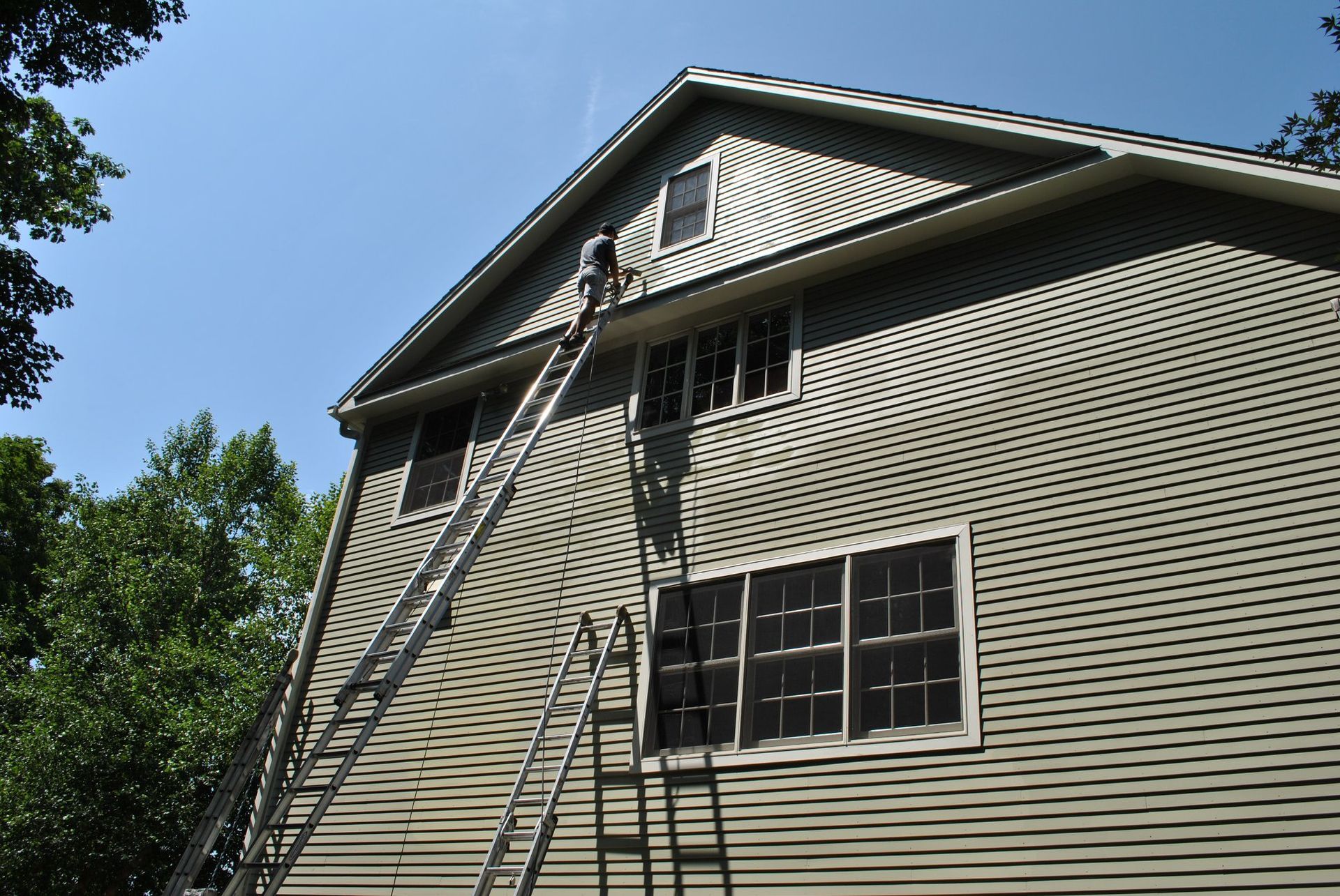 Person on ladder painting the side of a two-story house with green siding under a blue sky.