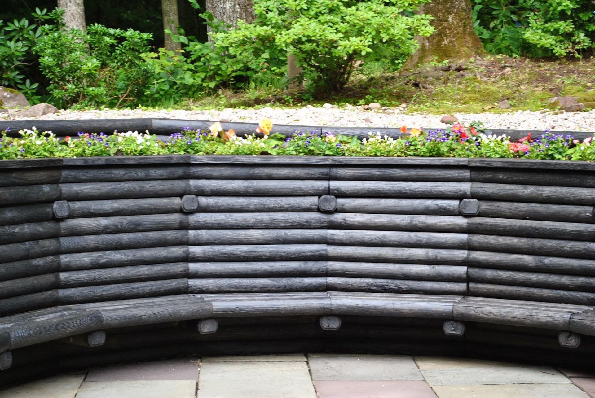 Curved wooden bench in a garden setting, with a flower bed behind it and trees in the background.