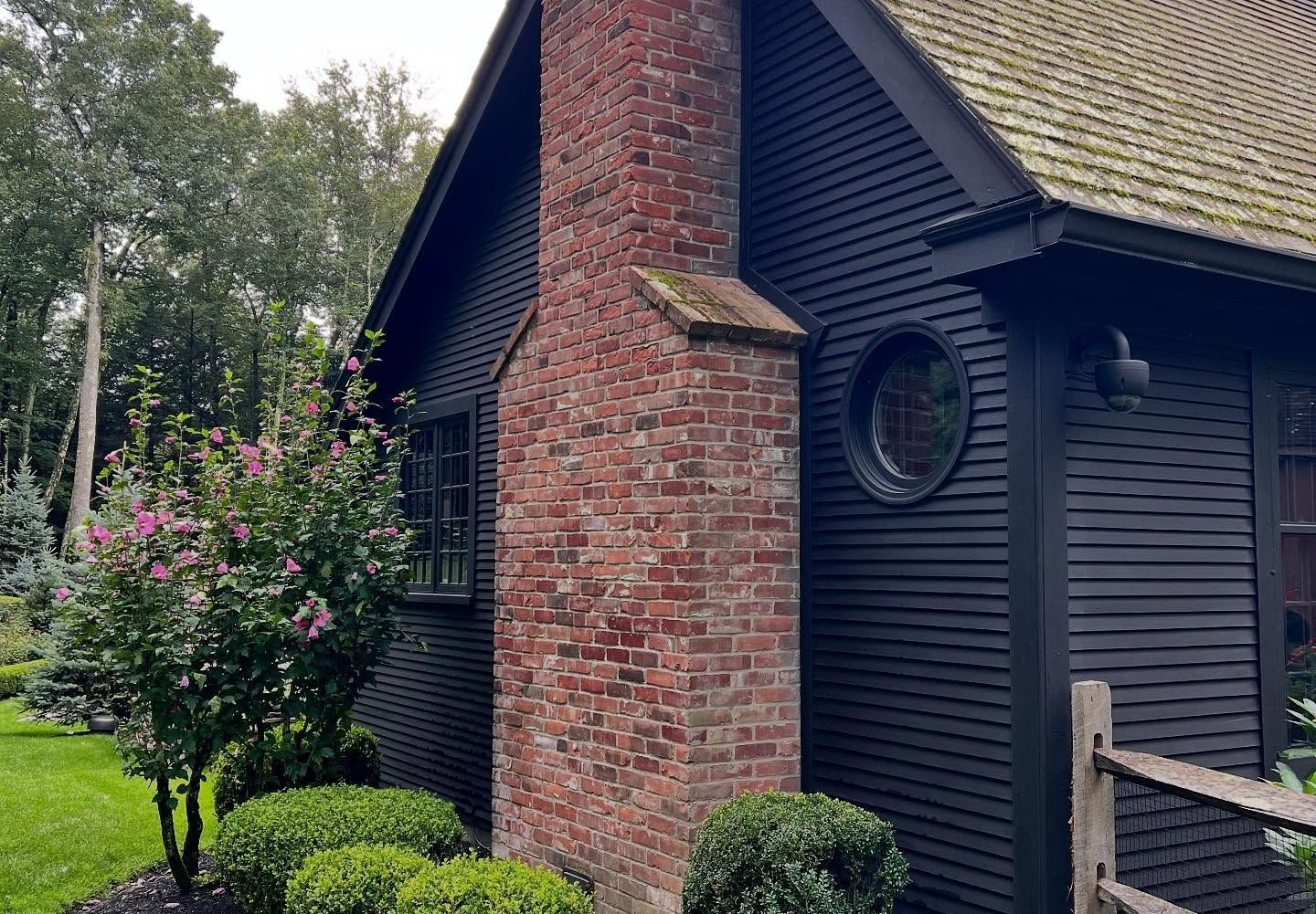 Black house with brick chimney, small round window, and shingled roof, surrounded by greenery.