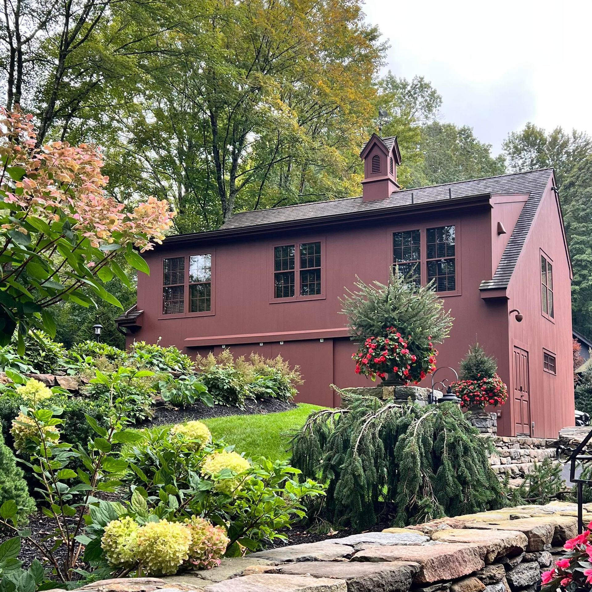 Red-painted building with windows, surrounded by lush green foliage and flowers, with a stone wall in the foreground.
