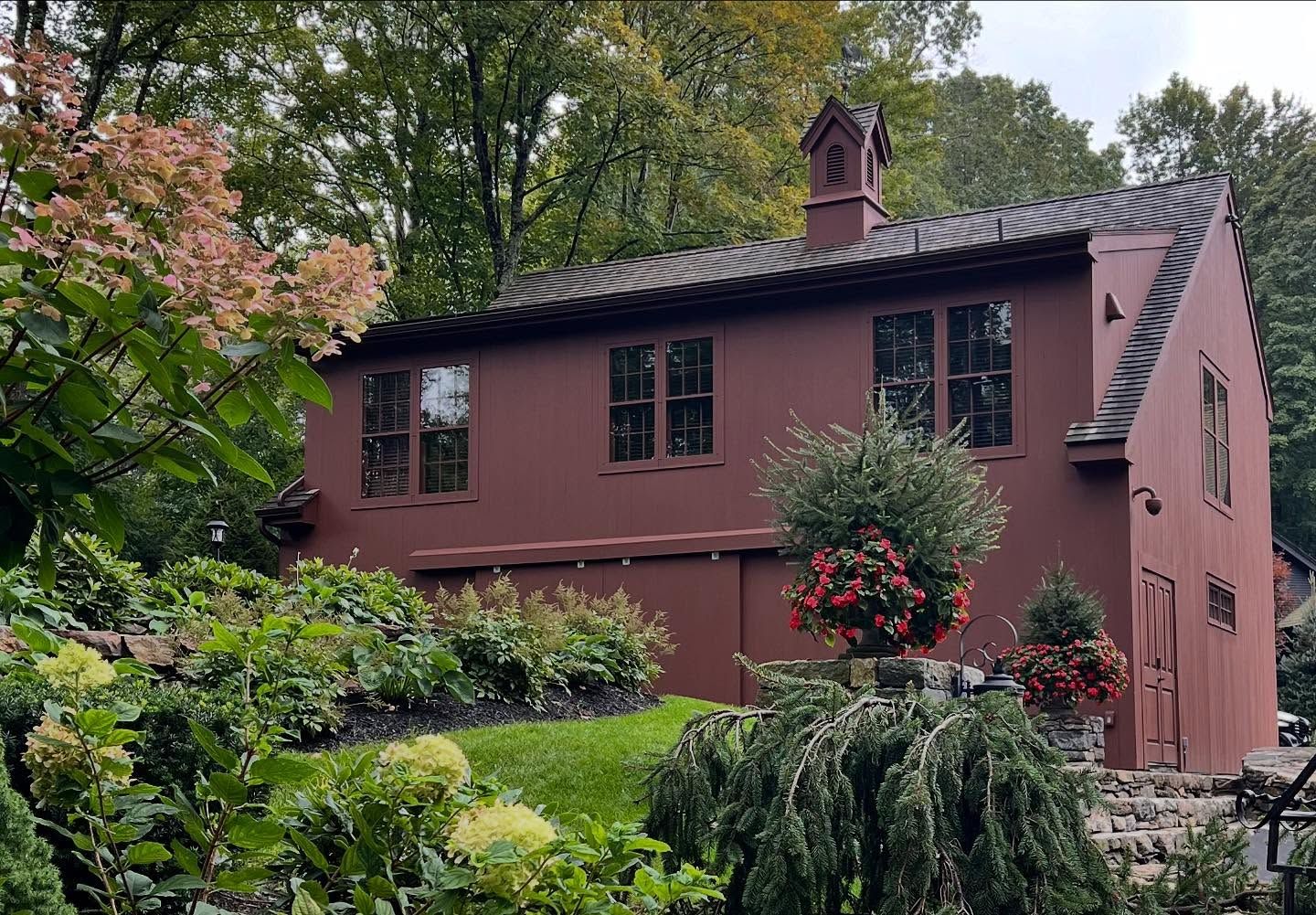 Maroon-colored house with dark roof and windows, nestled in a lush garden with flowers and greenery.