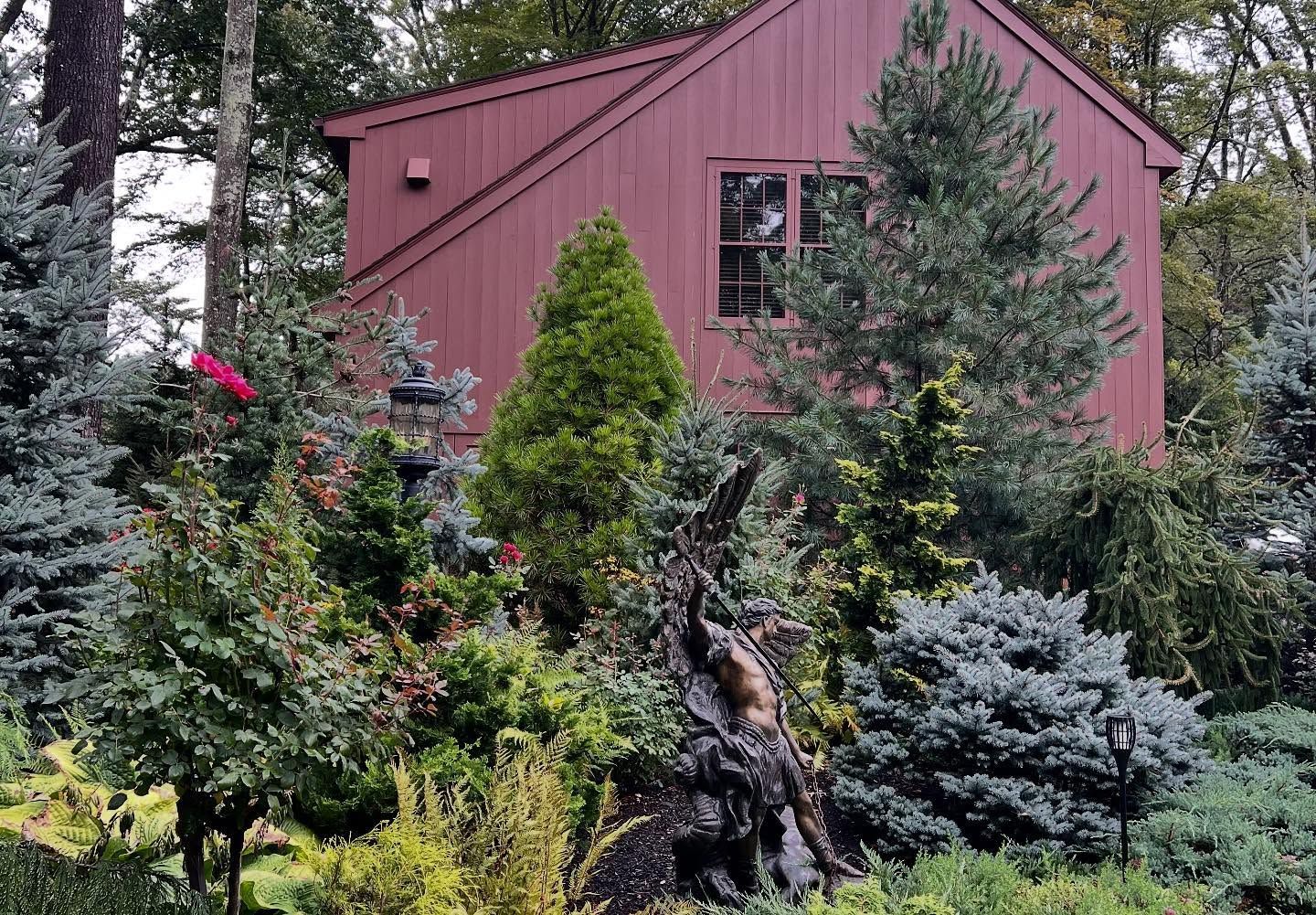 Red house surrounded by various green and blue-green evergreen trees and bushes.
