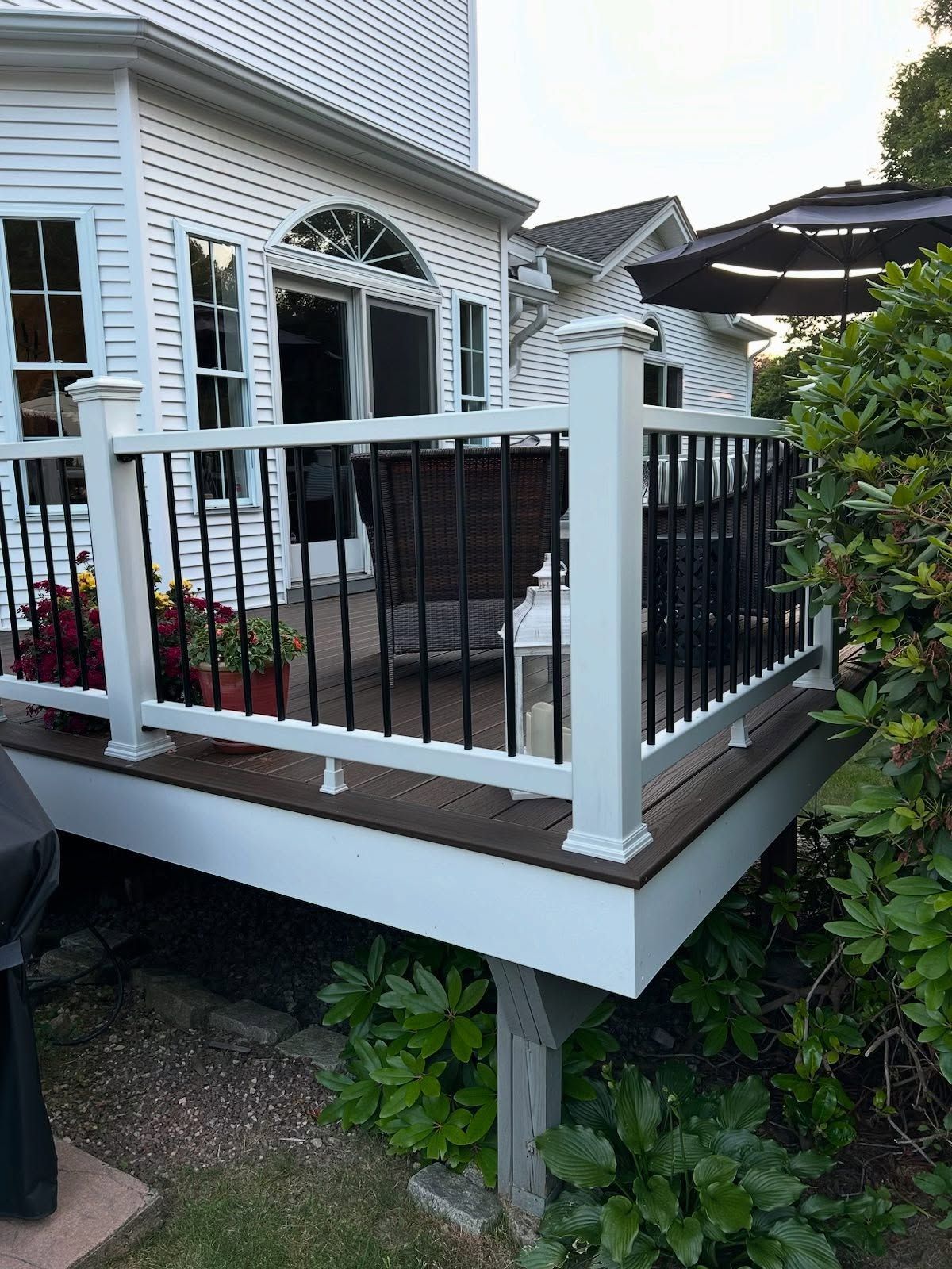 White house deck with white railings, black balusters, and brown decking. Plants and patio furniture are visible.