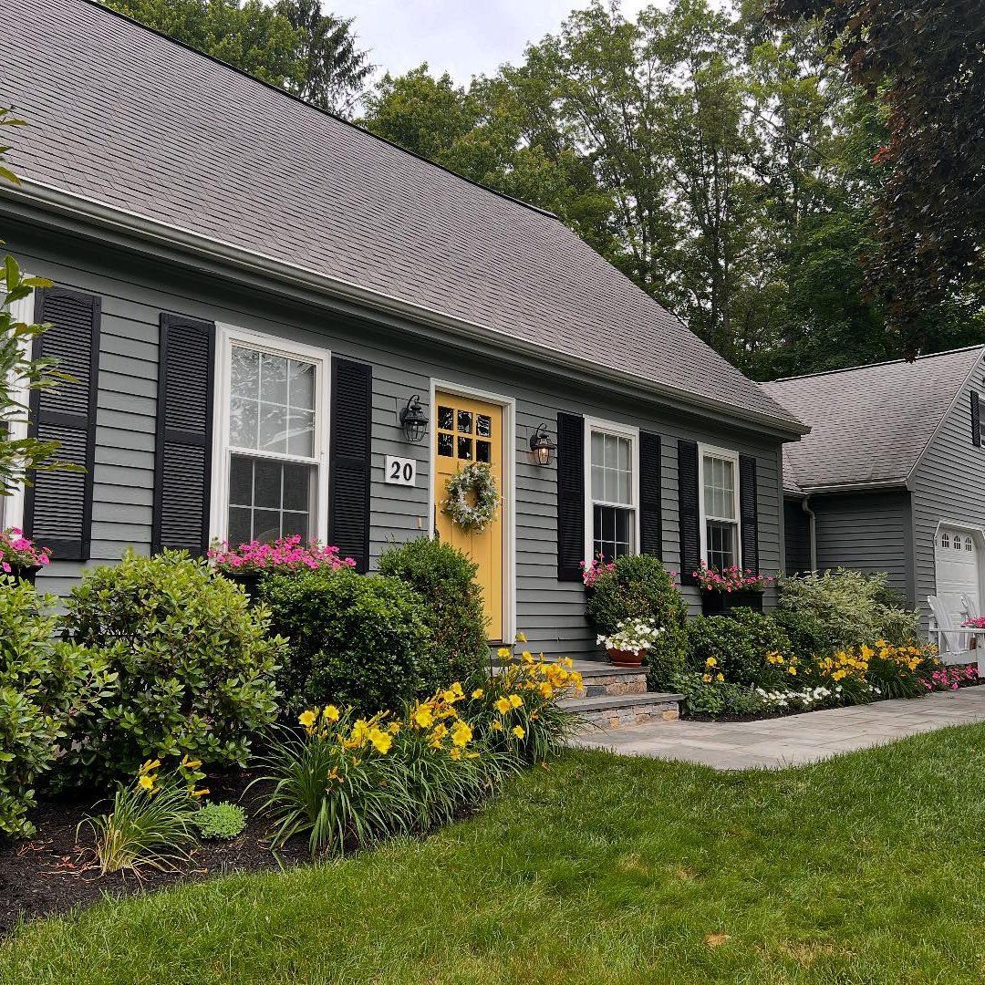 Gray house with black shutters, yellow door, and flower-filled garden.
