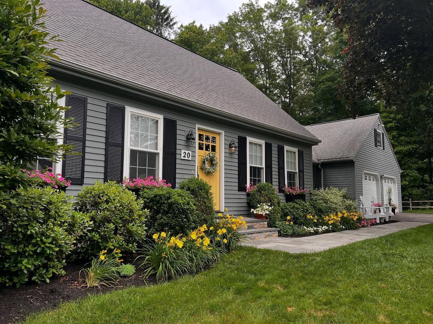 Gray house with yellow door, black shutters, and flower beds.