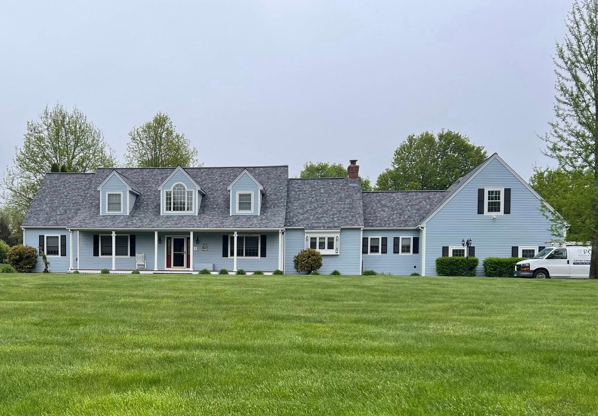 Blue house with green lawn, dormers, and a gray roof under a cloudy sky.