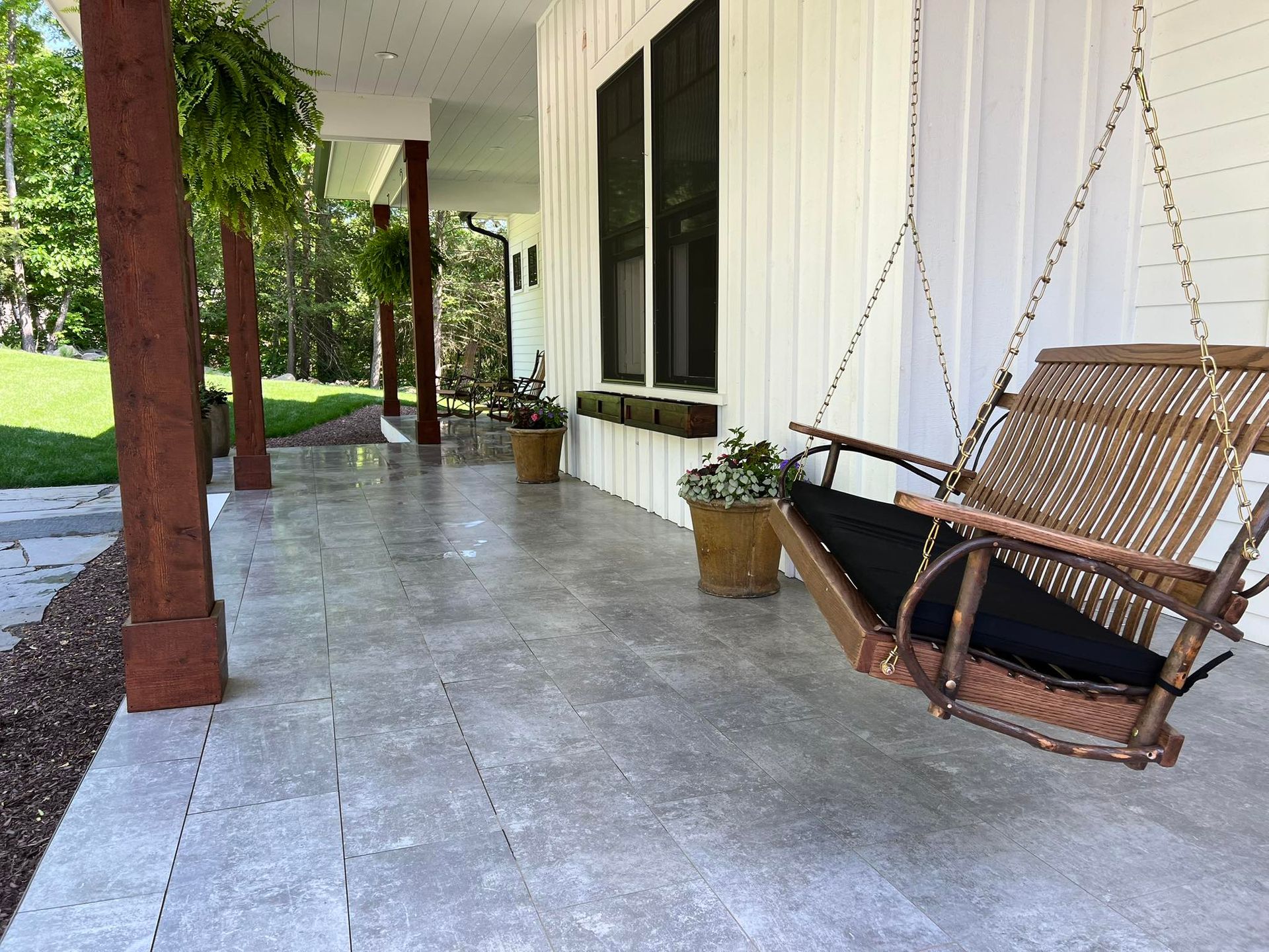 Porch with gray concrete floor, brown columns, a hanging wicker swing, and potted plants.