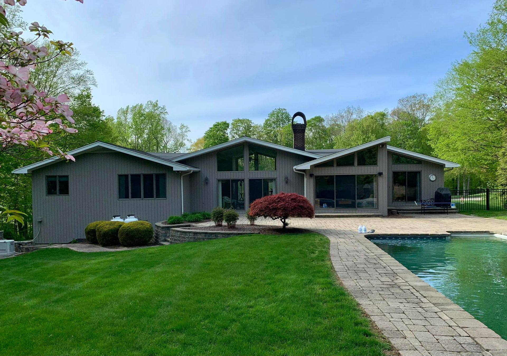 Gray house with a pool and lush greenery. Sunny day, with a red Japanese maple tree.
