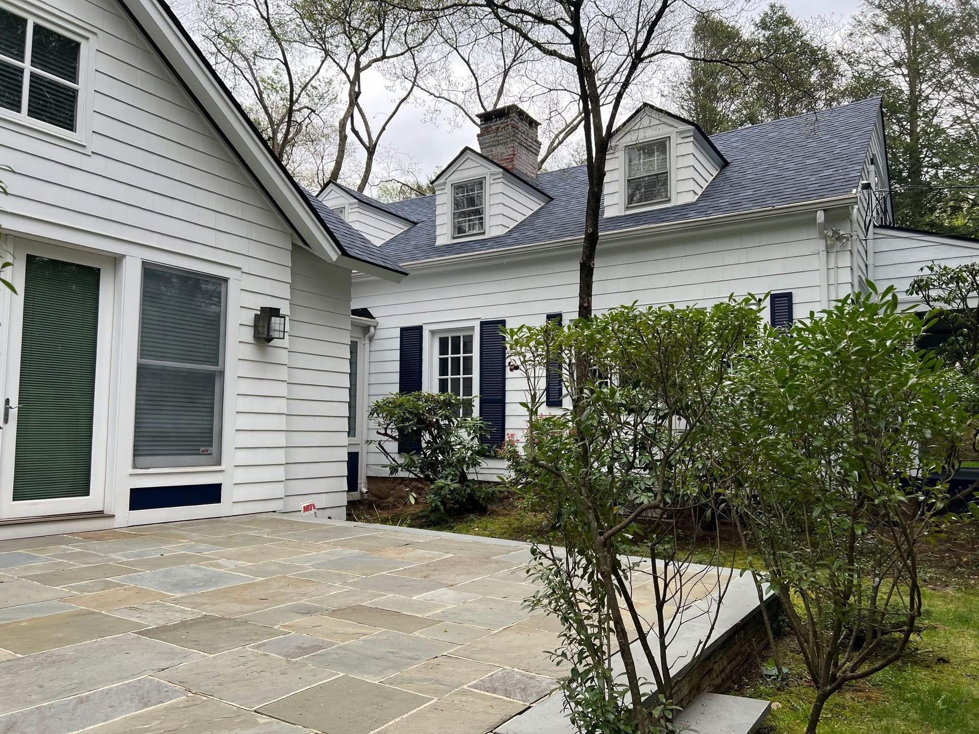 White house with blue shutters, slate patio, and lush green bushes.