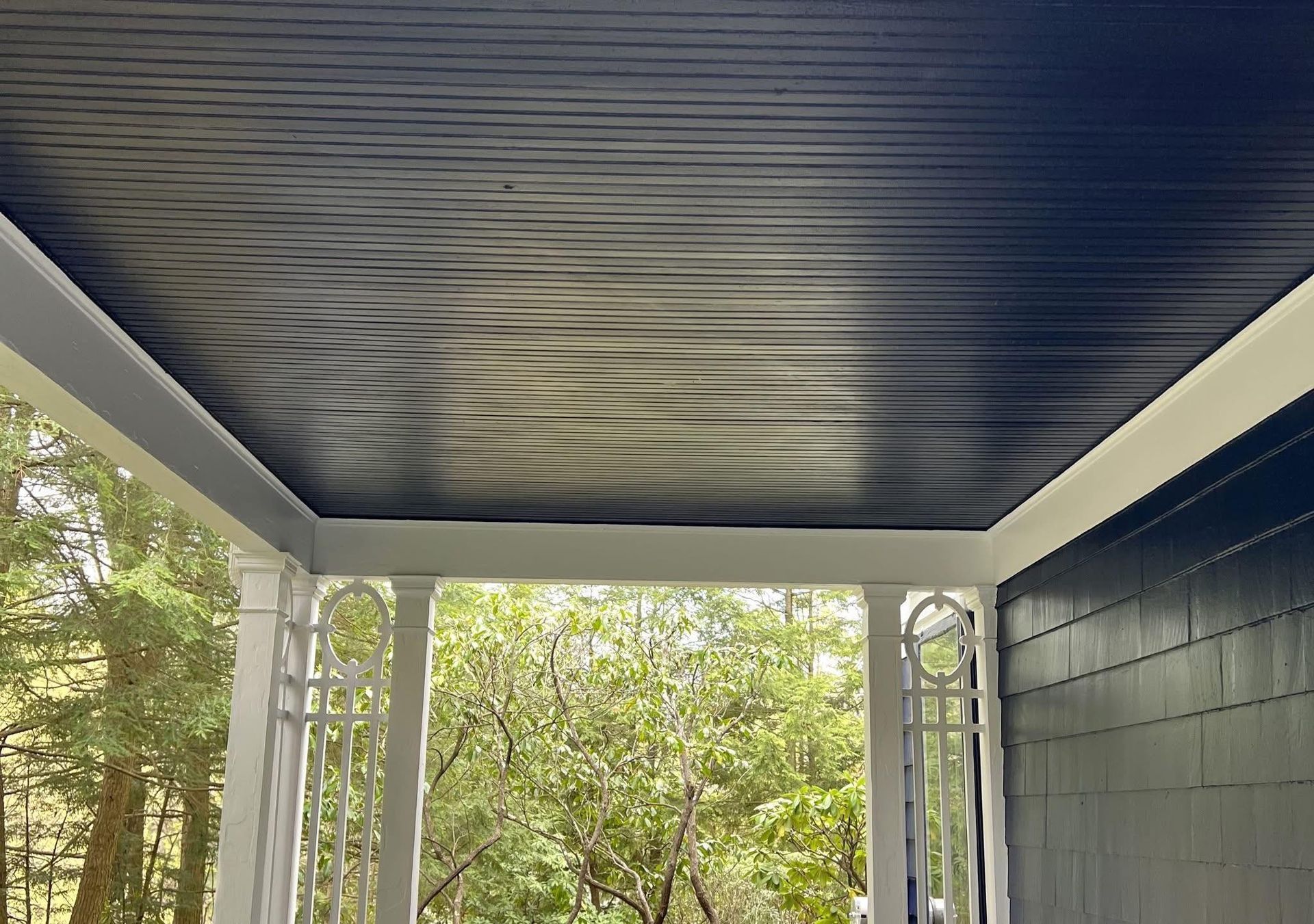 Porch ceiling with wood grain pattern, bordered by white trim. View includes foliage and building.