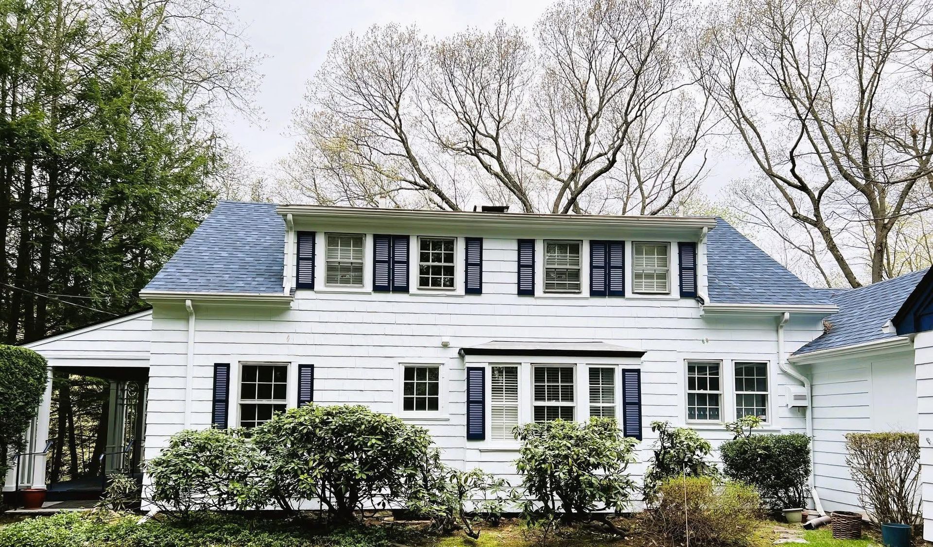 White two-story house with blue roof and shutters, surrounded by trees and bushes.