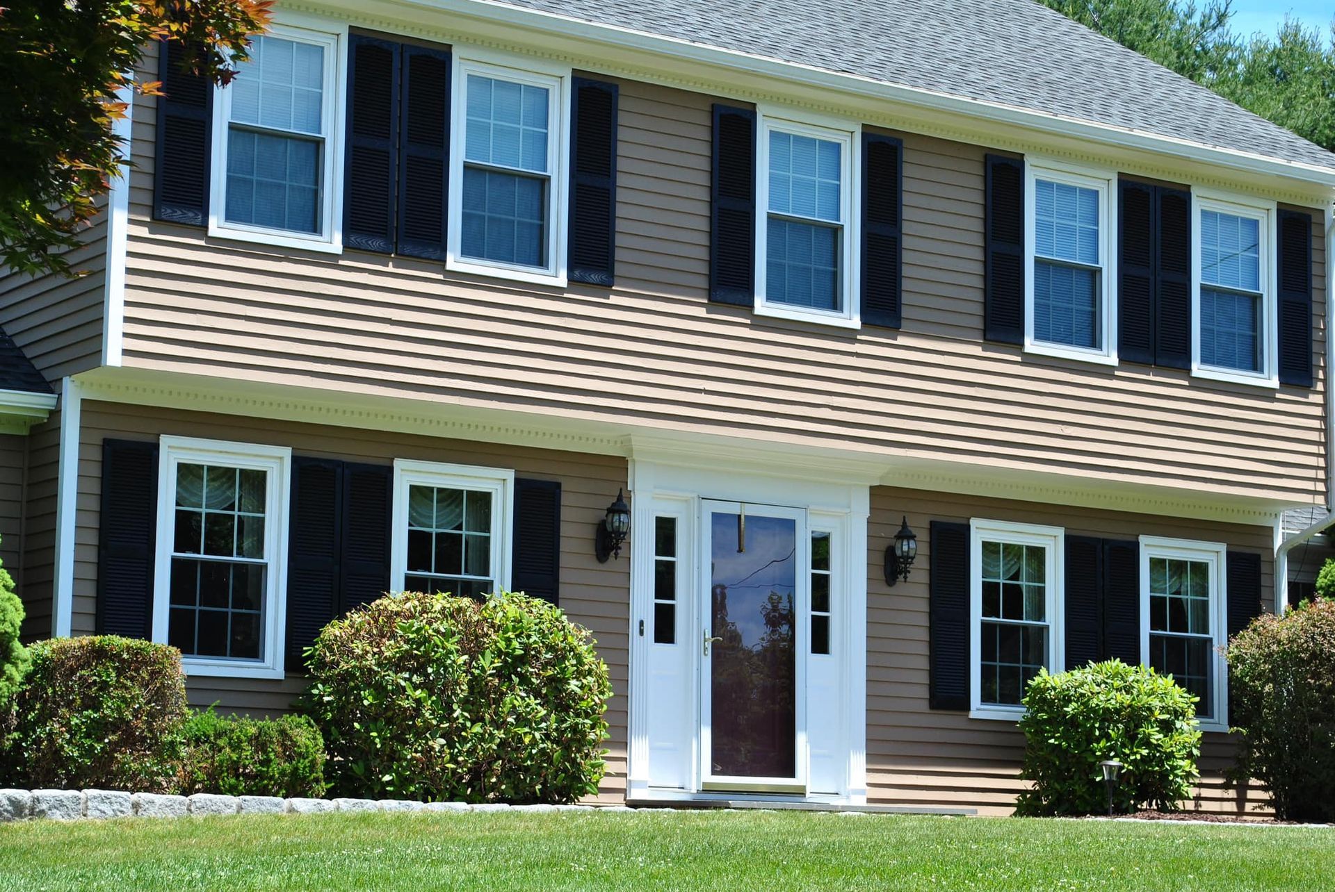 Two-story house with tan siding, black shutters, white trim, and a brown front door.
