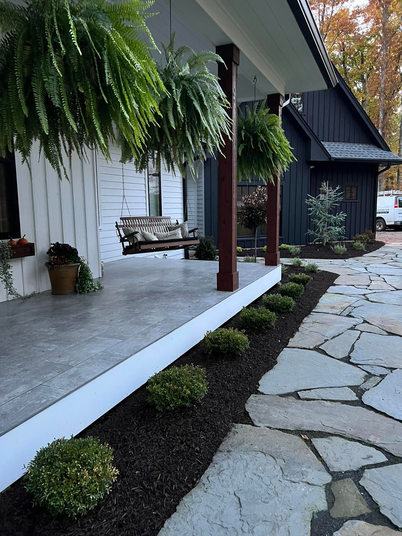 White porch with hanging ferns and a porch swing; black house in the background. Stone path.
