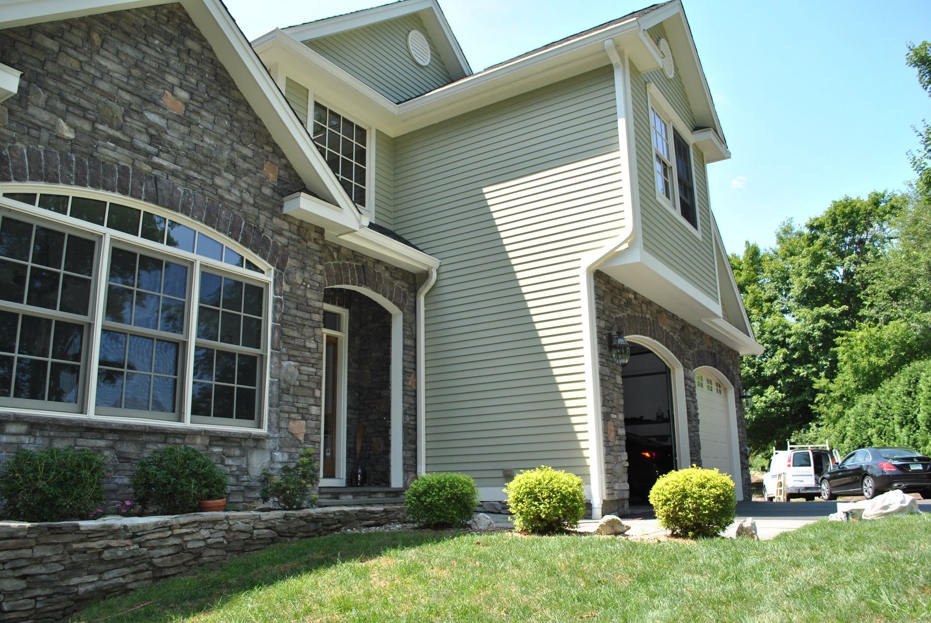 House exterior with stone and green siding, arched entry, garage, and landscaping on a sunny day.