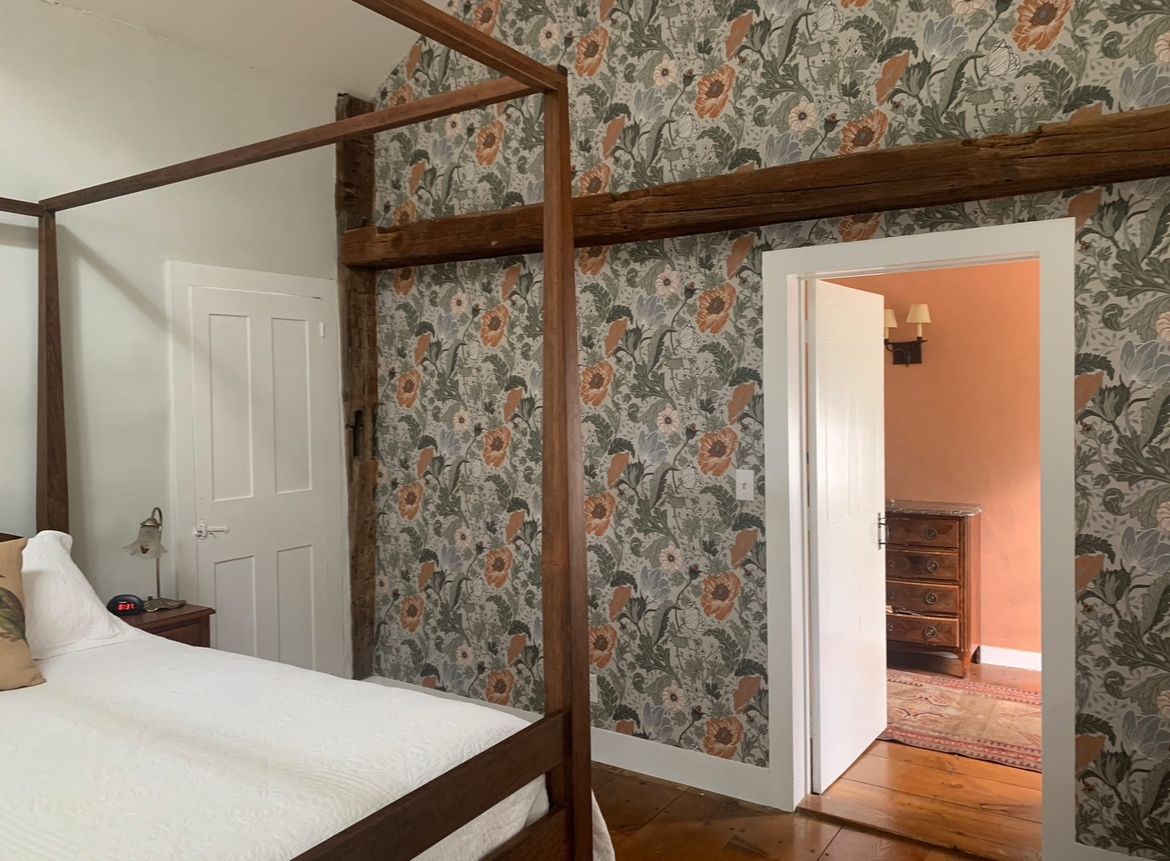 Bedroom with four-poster bed and floral wallpaper. An open door reveals a room with a dresser.