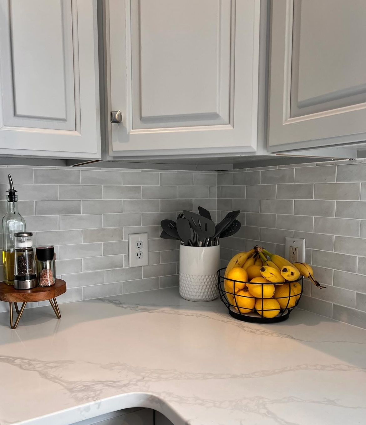 Kitchen countertop with gray cabinets, tiled backsplash, and white marble-look countertop. Fruit basket of lemons, utensils, and spice rack.