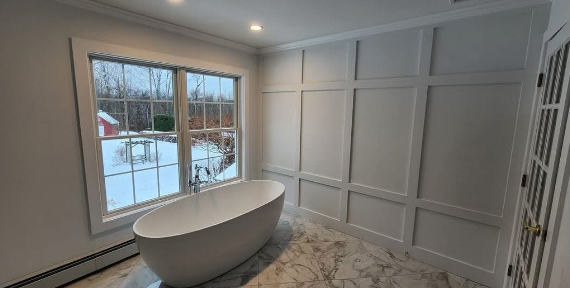 White bathroom with a freestanding tub by a window overlooking a snowy yard. White paneling covers the wall.