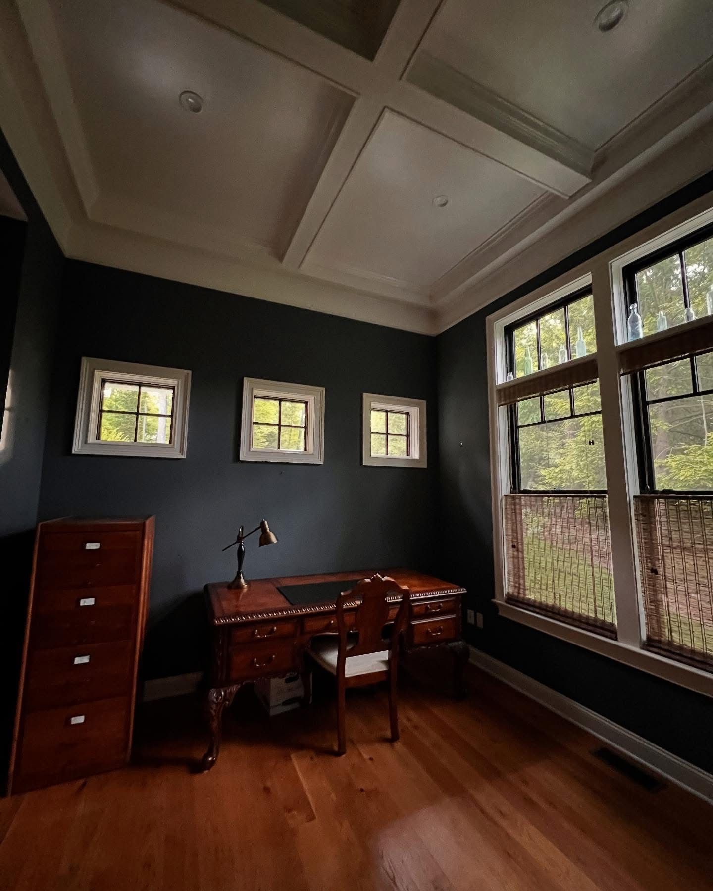 Dark blue home office with wood desk, cabinets, and hardwood floors. Three small windows line the left wall, two large windows on the right.