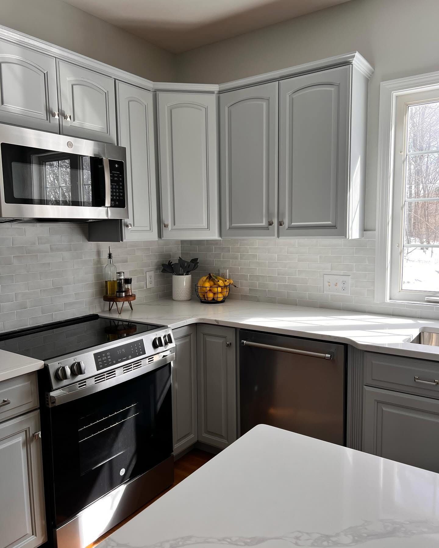 A modern kitchen with gray cabinets, white countertops, stainless steel appliances, and a tiled backsplash. Sunlight streams in from a nearby window.